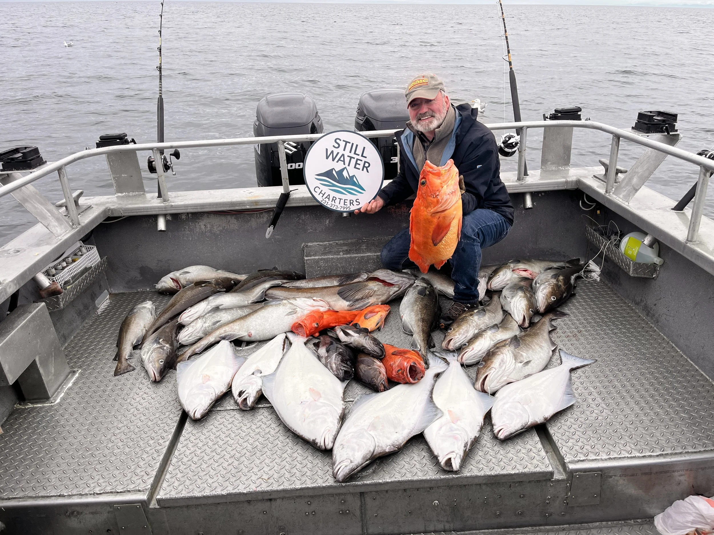 Man holding an orange fish on boat surrounded by multiple caught fish, with a sign that reads 'Still Water Charters'.