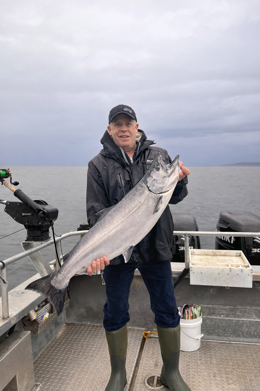 A man smiling on a boat holding a large fish he caught, with a body of water and cloudy sky in the background, wearing rubber boots and a dark jacket.