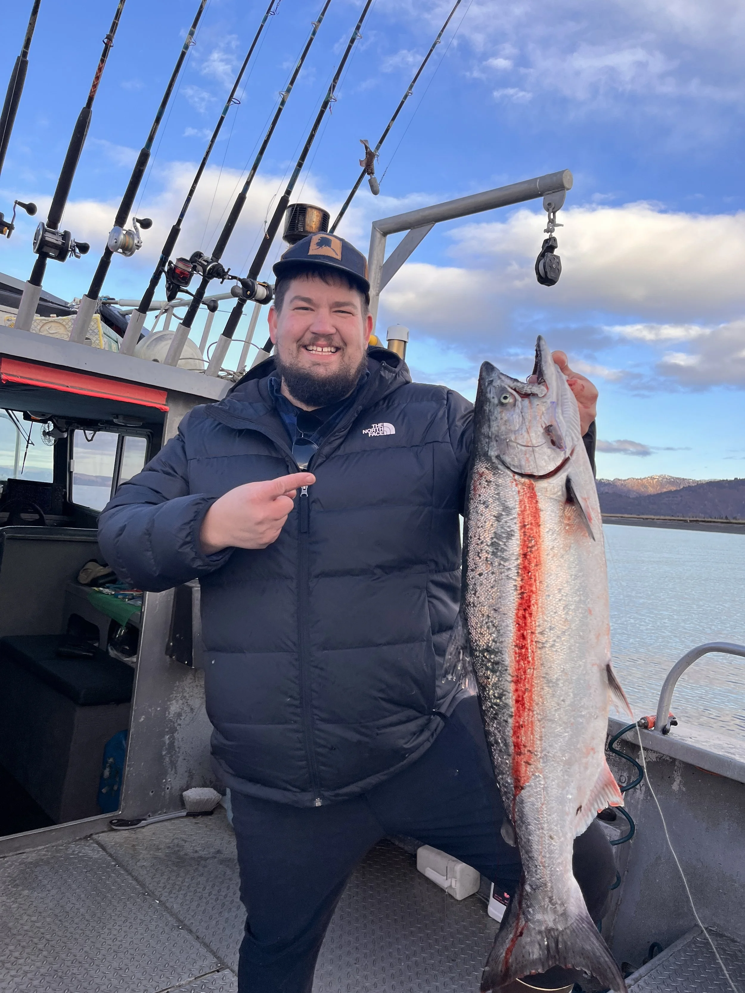 Man smiling and pointing at a large fish he caught on a boat, with fishing rods mounted in the background and water in the distance.