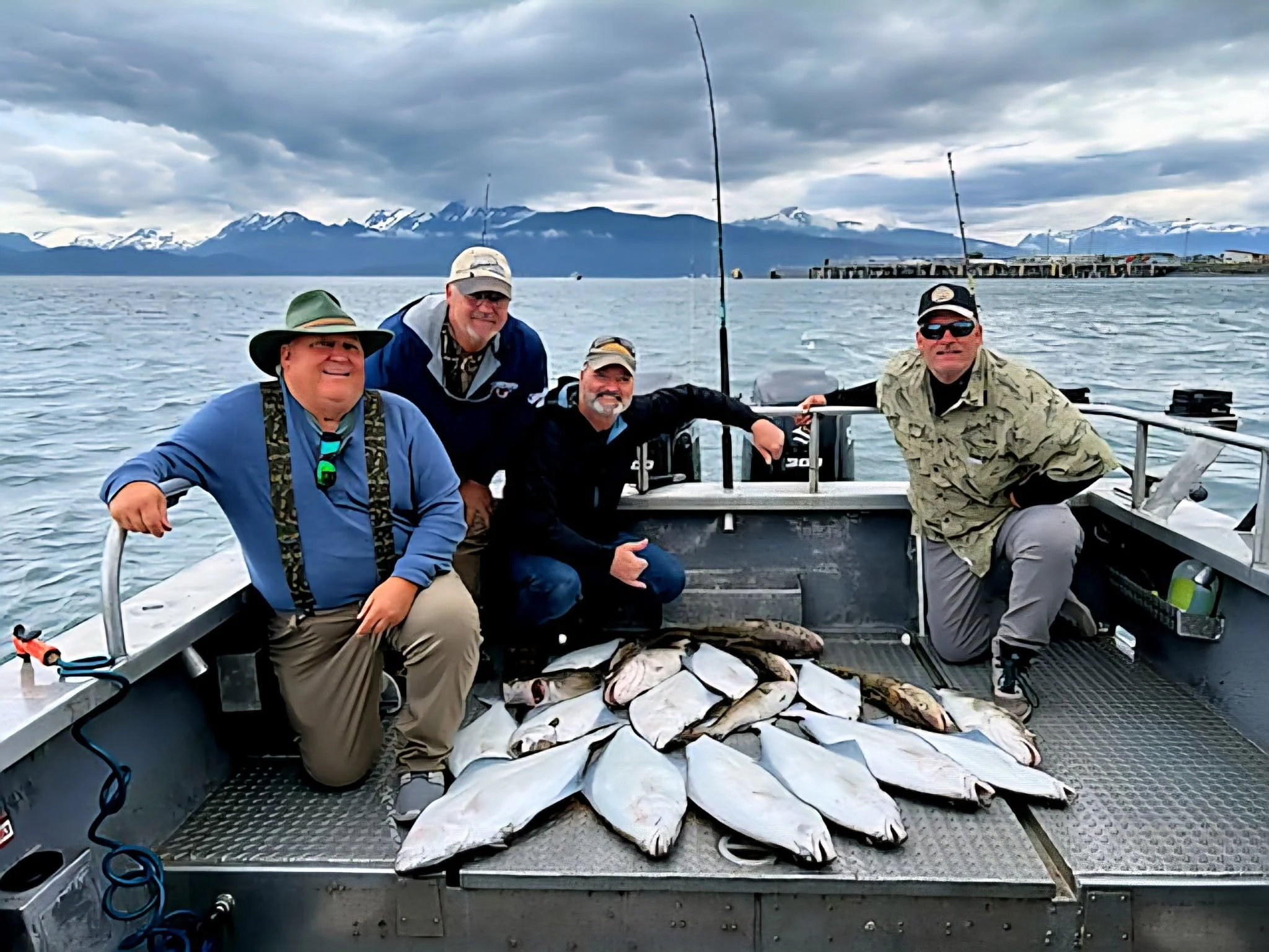 Four men on a boat with a catch of fish, with mountains and water in the background.