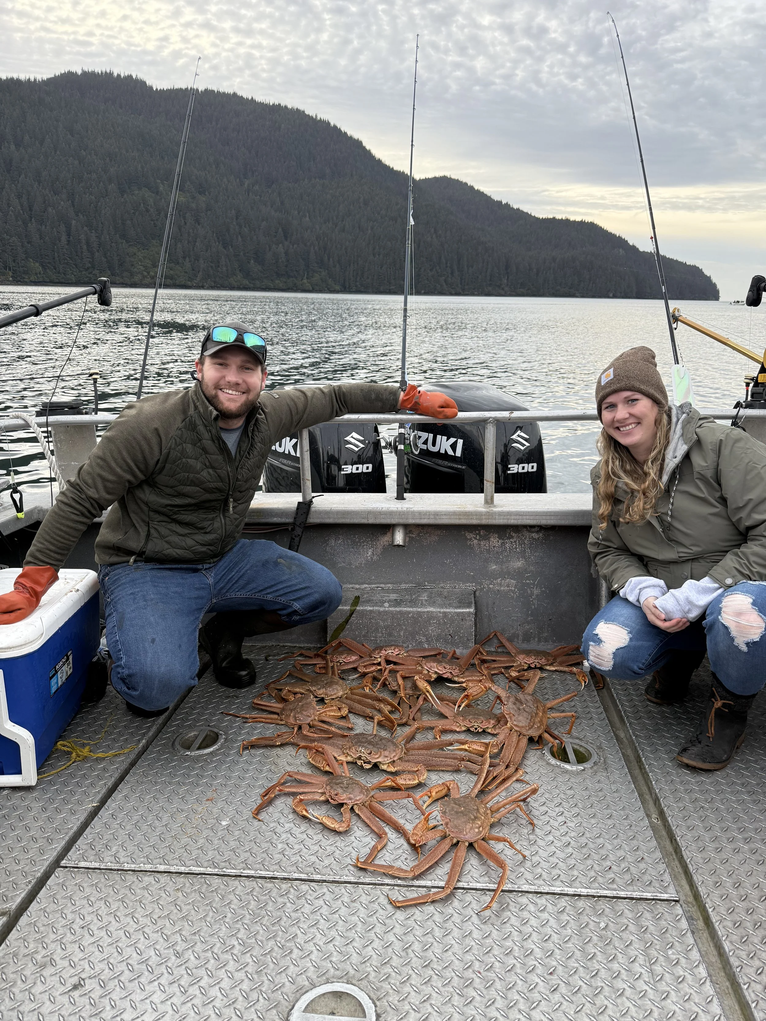 Two people on a boat displaying a catch of crabs, with fishing rods in the background and a mountainous landscape and water behind them.