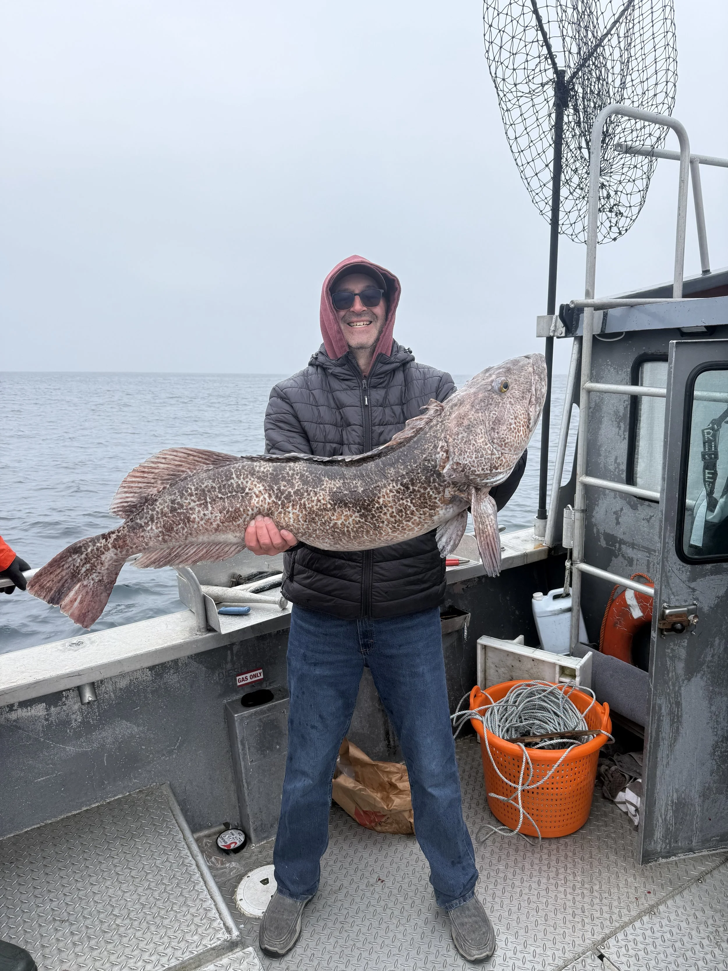 Man wearing sunglasses, a hooded jacket, and jeans holding a large fish on a boat in the water.