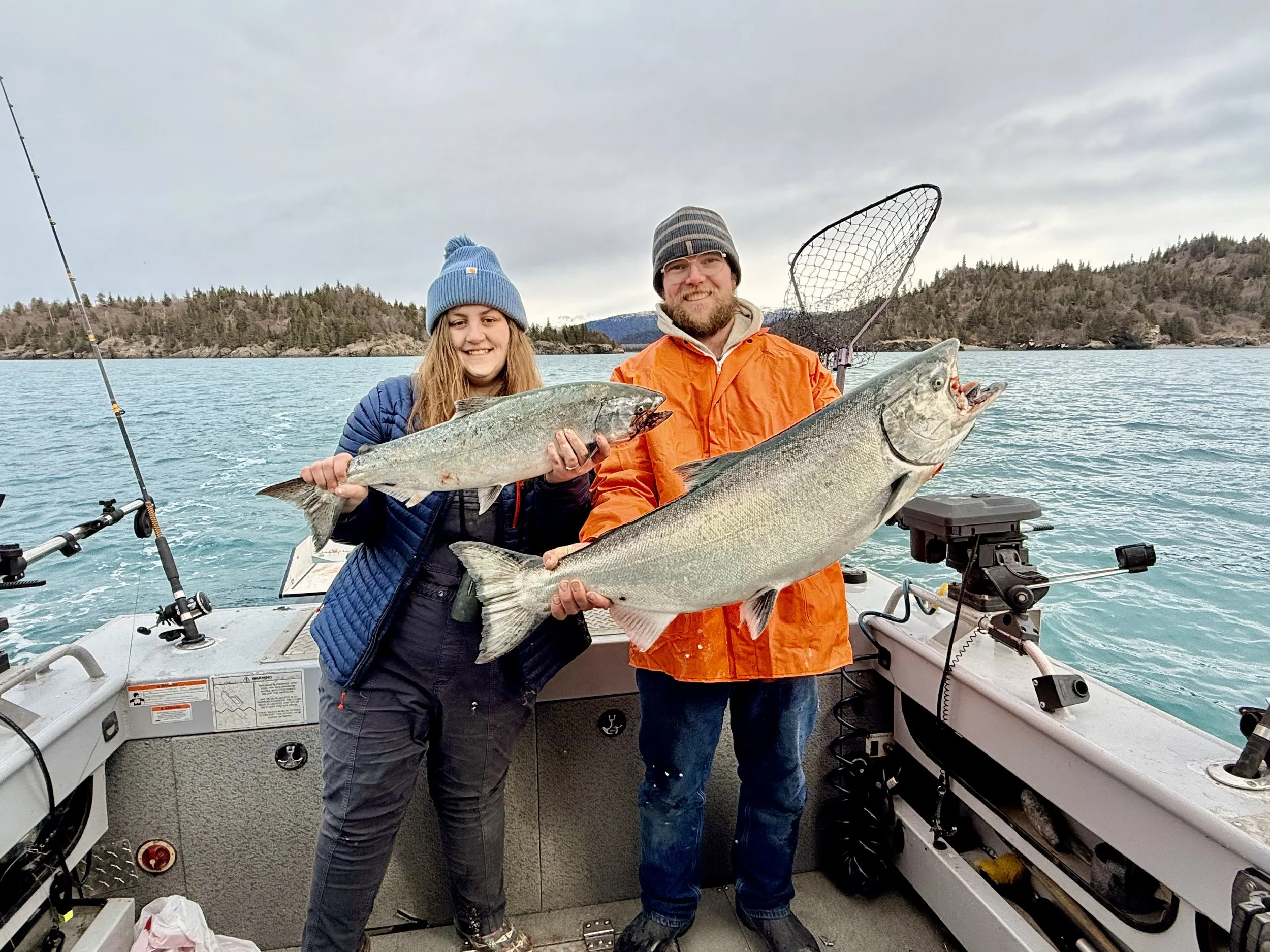 Two people on a boat holding large fish they caught, with water and land in the background.