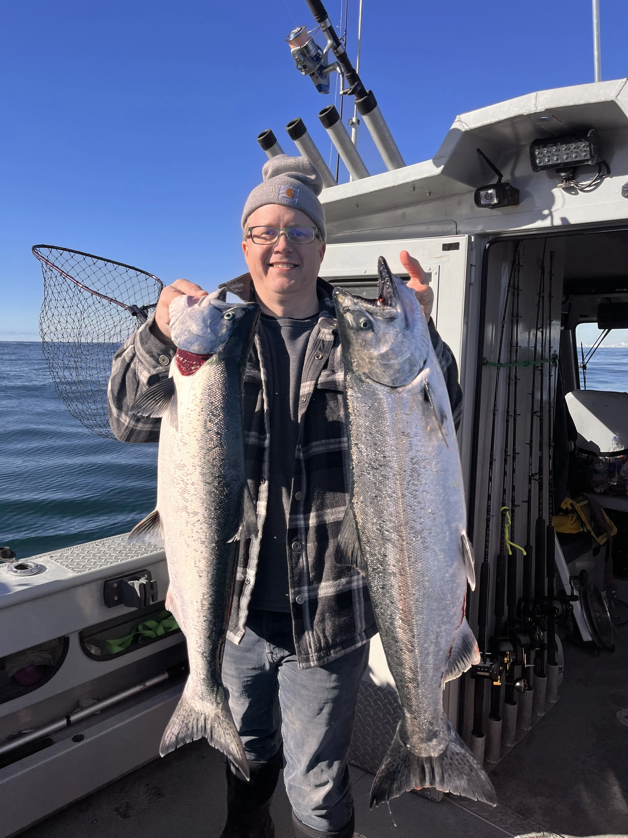 Man on a boat holding two large fish, blue sky, fishing gear visible.