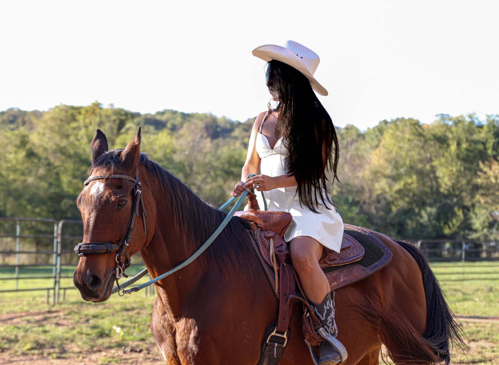 A woman with long black hair wearing a white cowboy hat and white dress riding a brown horse in a field with trees in the background.