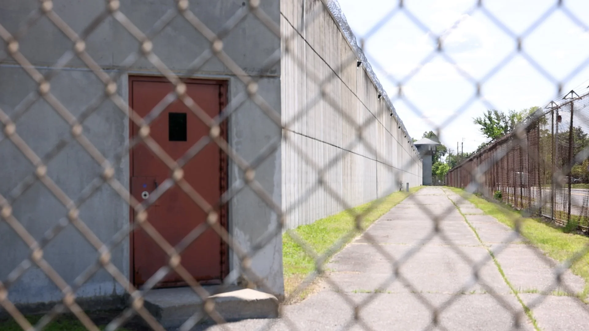 View through a chain-link fence showing MCI-Concord, a now-closed prison in Massachusetts. Photo shows red metal door on the left and a long alleyway extending to the right.