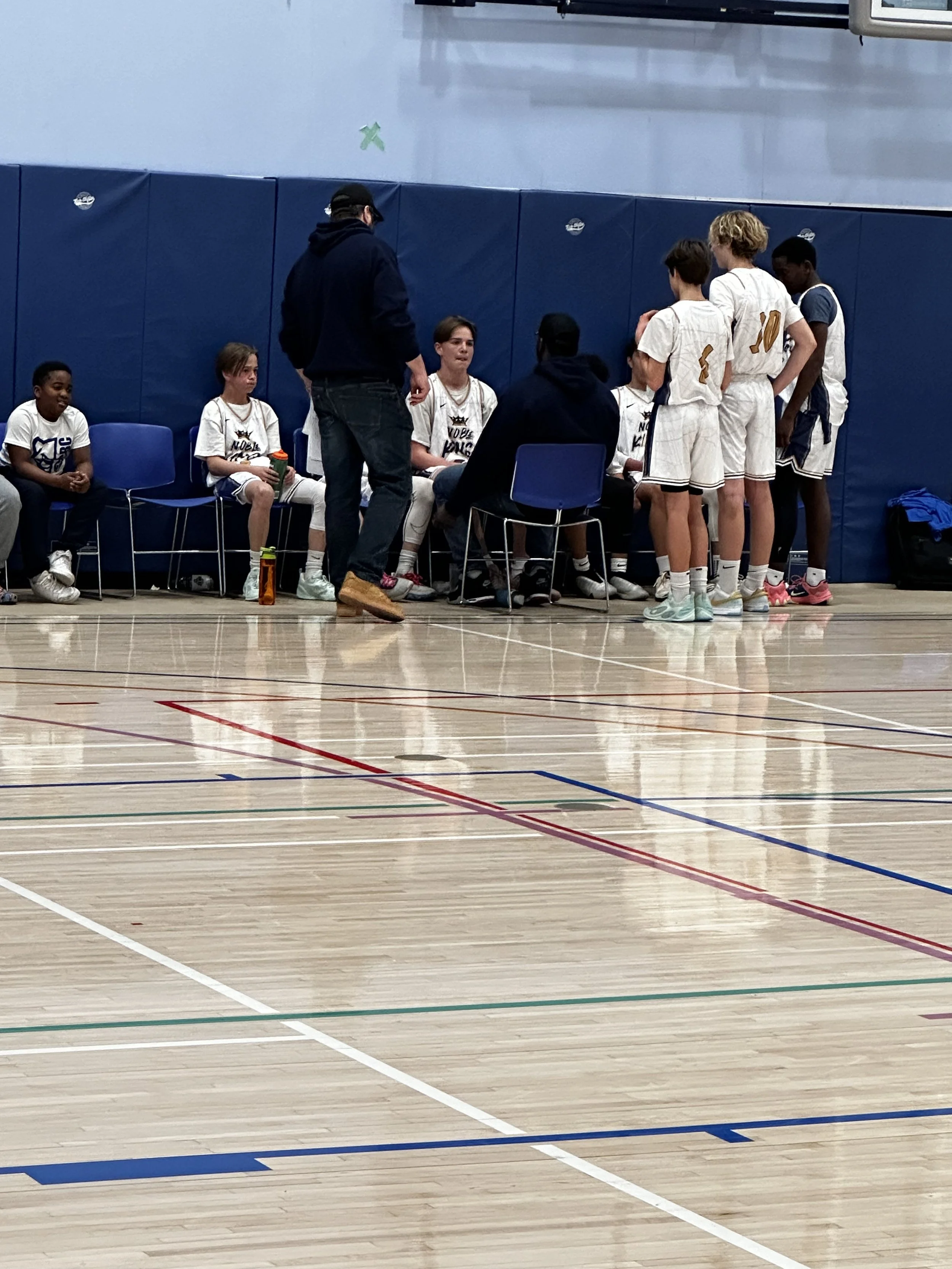 Youth basketball team huddle during a timeout in an indoor gym, with players sitting and standing, listening to coaches.