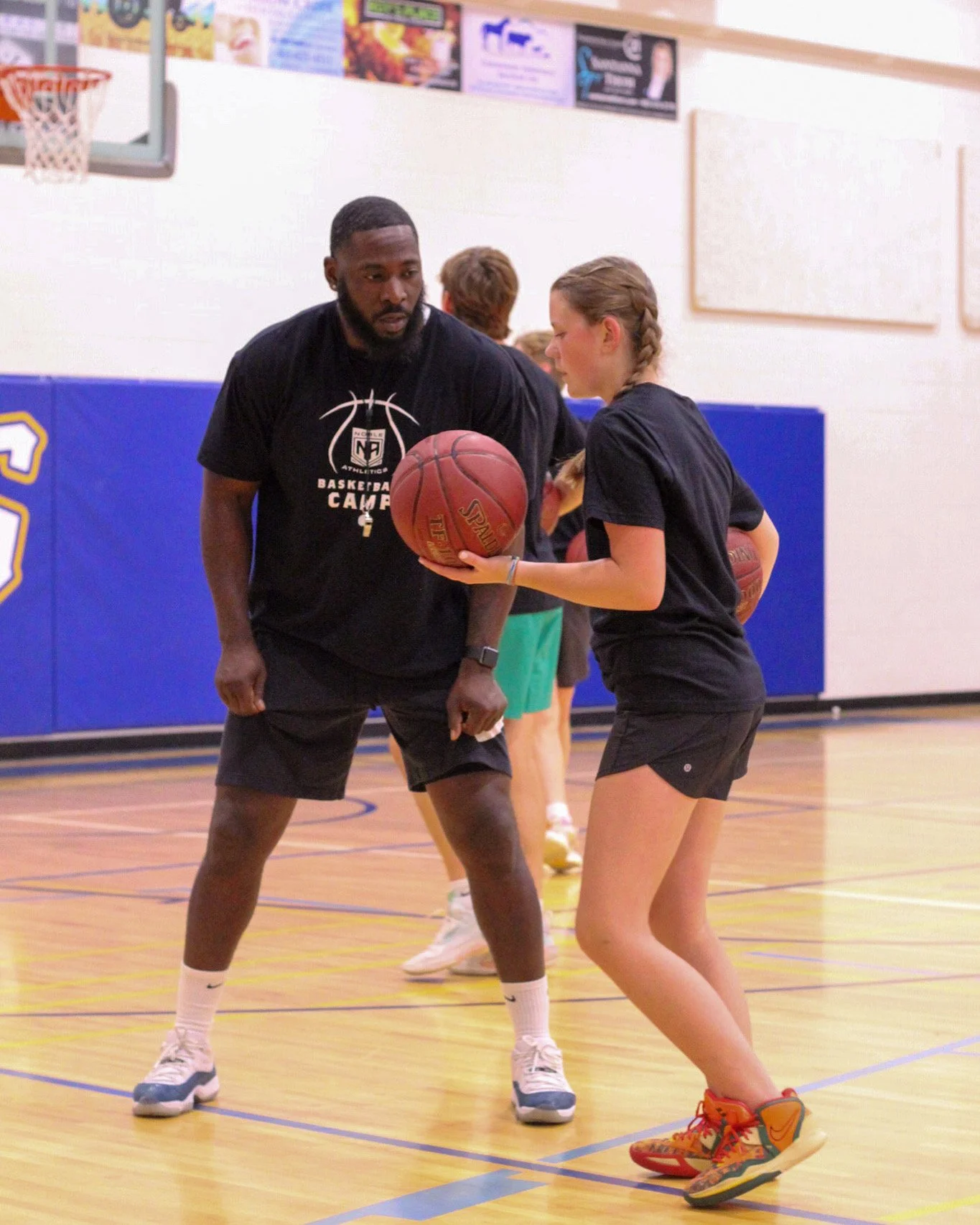 A coach instructs a young girl during a basketball practice in an indoor gymnasium. The girl is holding a basketball and wearing colorful athletic shoes. Other children are practicing in the background.