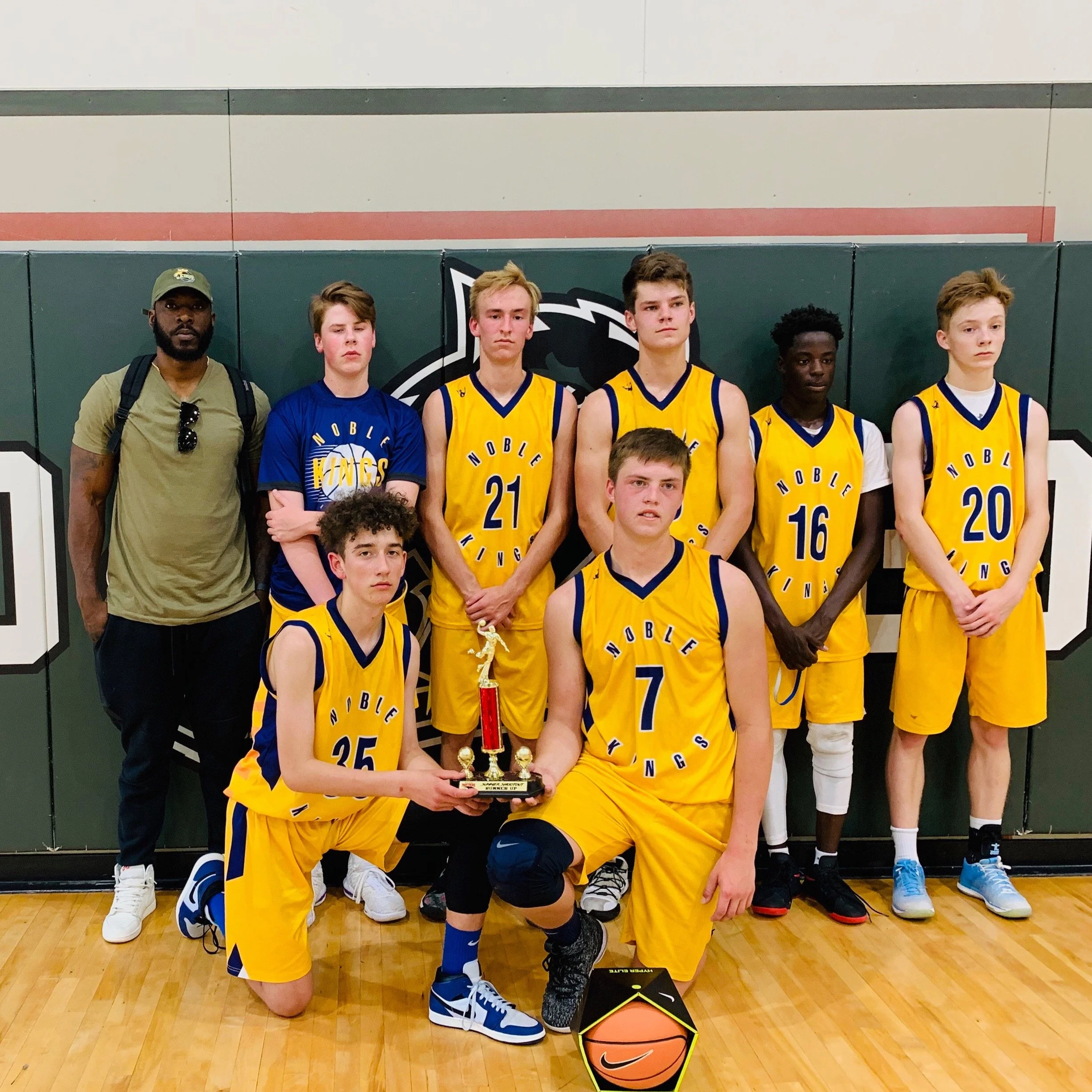 A youth basketball team posing indoors with a trophy, some team members kneeling, holding the trophy, wearing yellow and blue uniforms, on a basketball court.