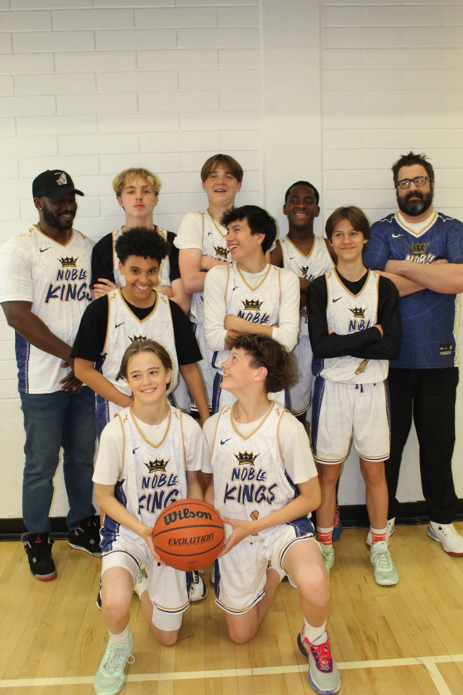 Group of young basketball players and coaches posing for a team photo indoors, with one player holding an orange Wilson basketball, all wearing white and black jerseys with 'Noble Kings' written on them.