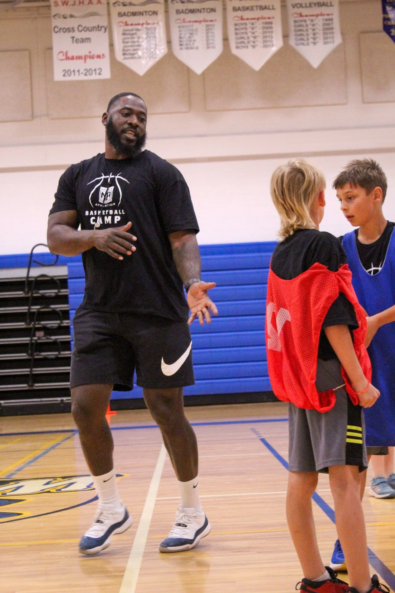 A coach and two young basketball players standing on an indoor basketball court, with championship banners hanging on the wall behind them.