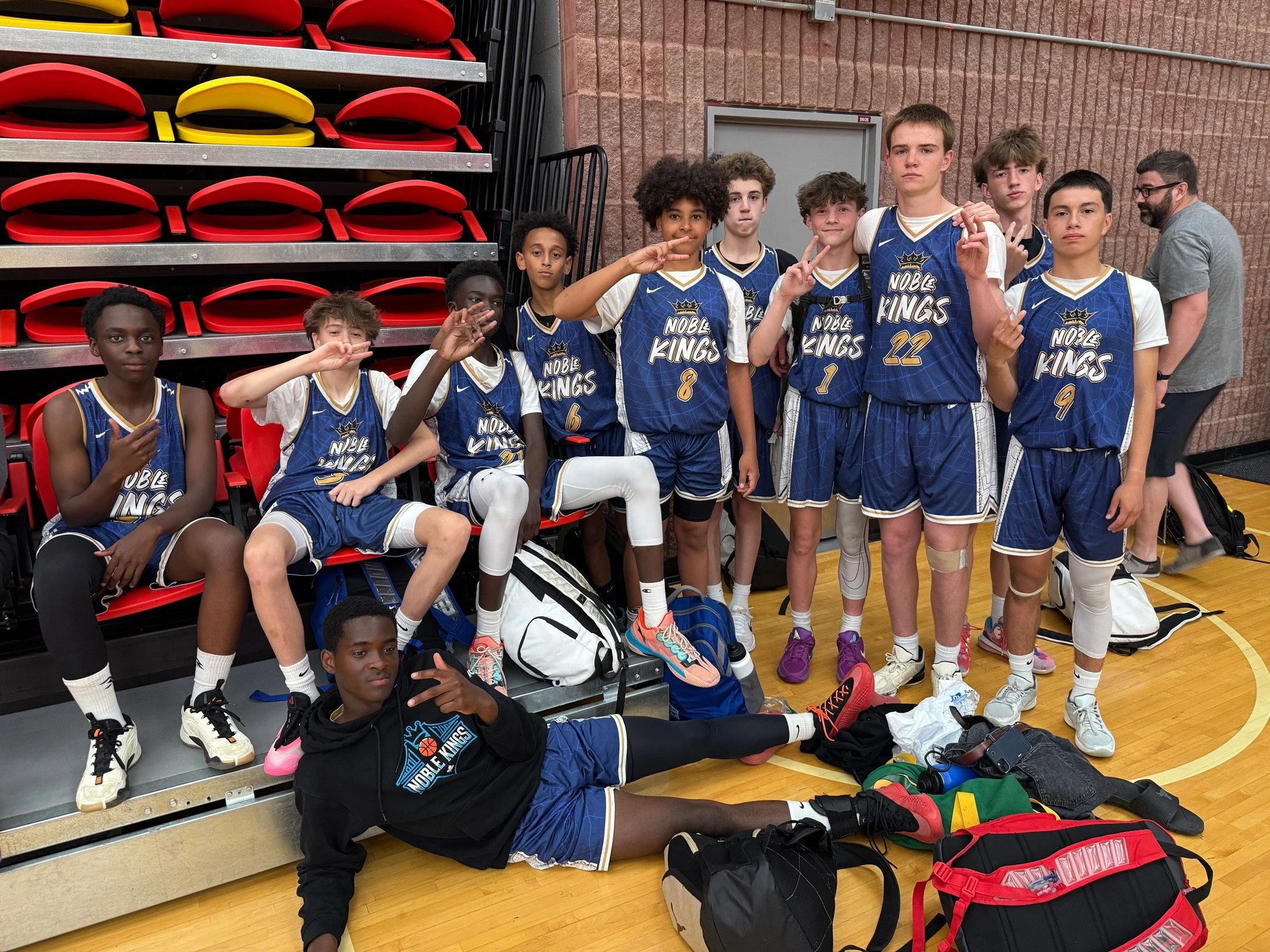 A group of young basketball players in blue jerseys with 'Noble Kings' on them, posing for a team photo in a gymnasium. They are making peace signs and sitting or standing near red bleachers with equipment bags on the floor.