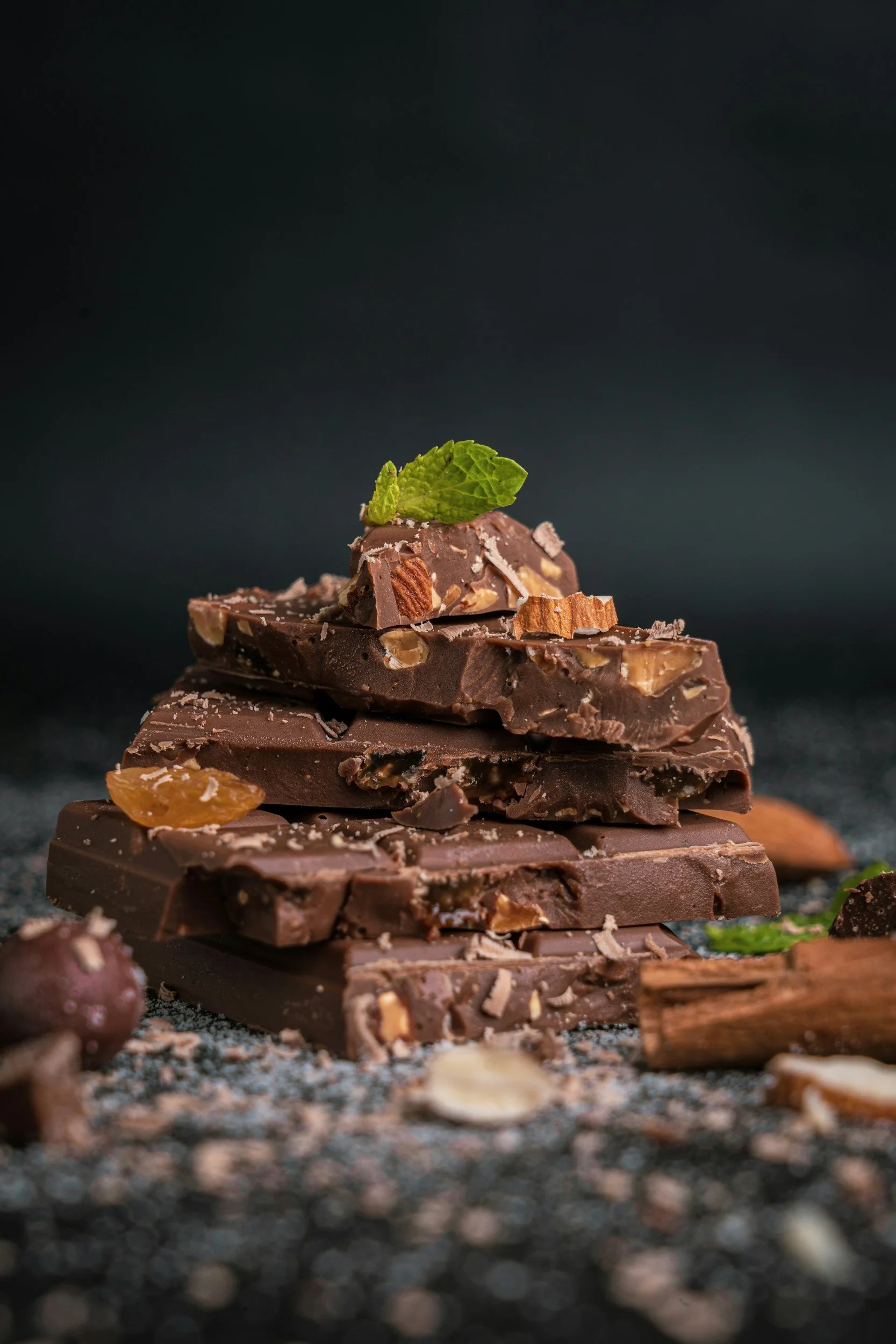 Stacked pieces of dark chocolate with nuts, topped with a mint leaf, surrounded by cinnamon sticks, almonds, and hazelnuts.