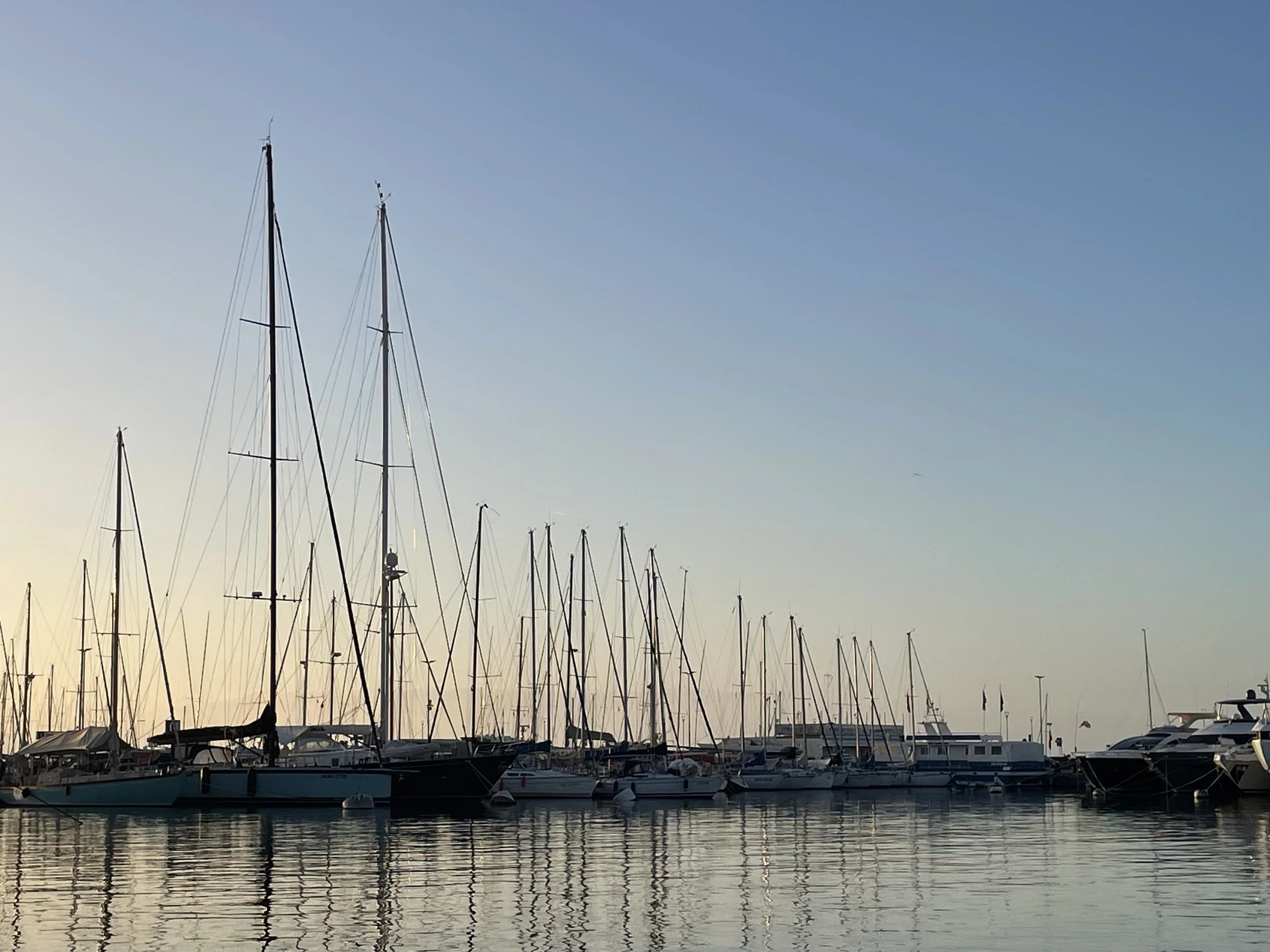 A marina with sailboats and yachts docked on calm water during sunset, with clear sky.