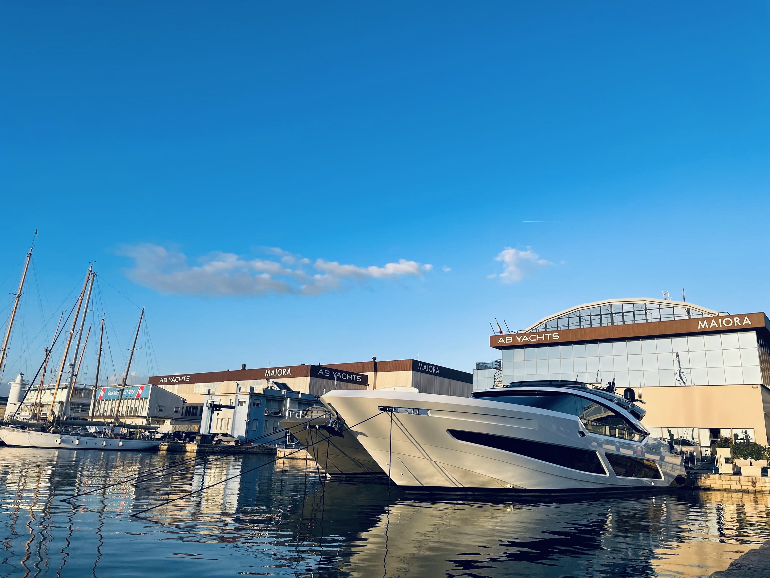Luxury yacht docked at a marina with several sailboats, modern buildings labeled 'AB YACHTS' and 'MAIORA', under a clear blue sky with a few scattered clouds.