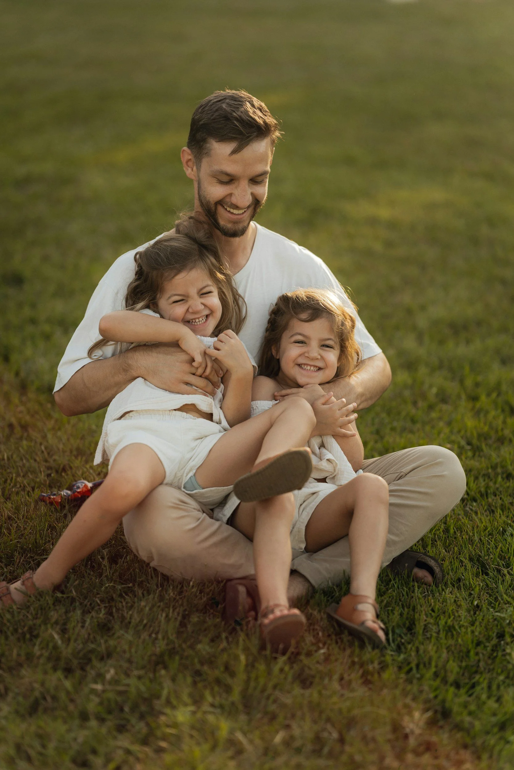 A man sitting on grass with two young girls, all smiling and hugging outdoors.