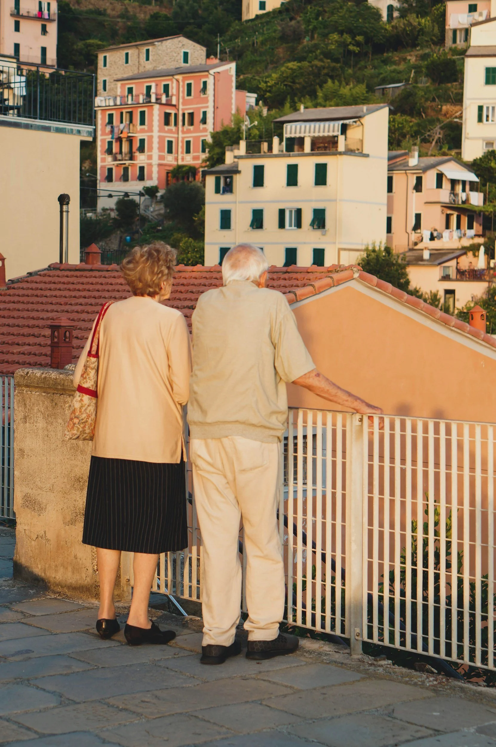 An elderly couple walking on a sidewalk in front of a white metal fence, with colorful buildings and green hills in the background, during sunset.