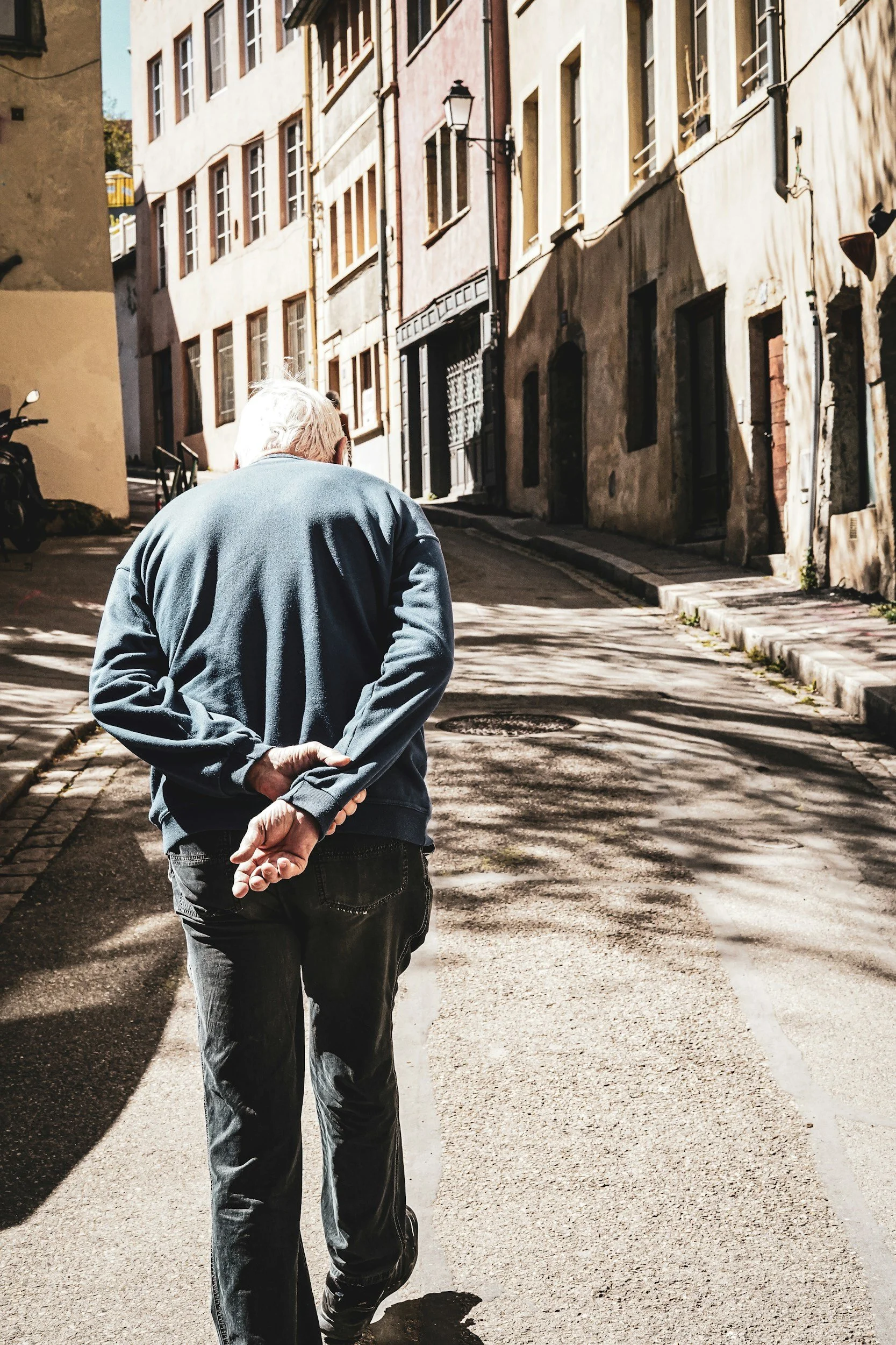 Older man with gray hair walking down a narrow, sunlit urban street with buildings on both sides, holding his hands behind his back.