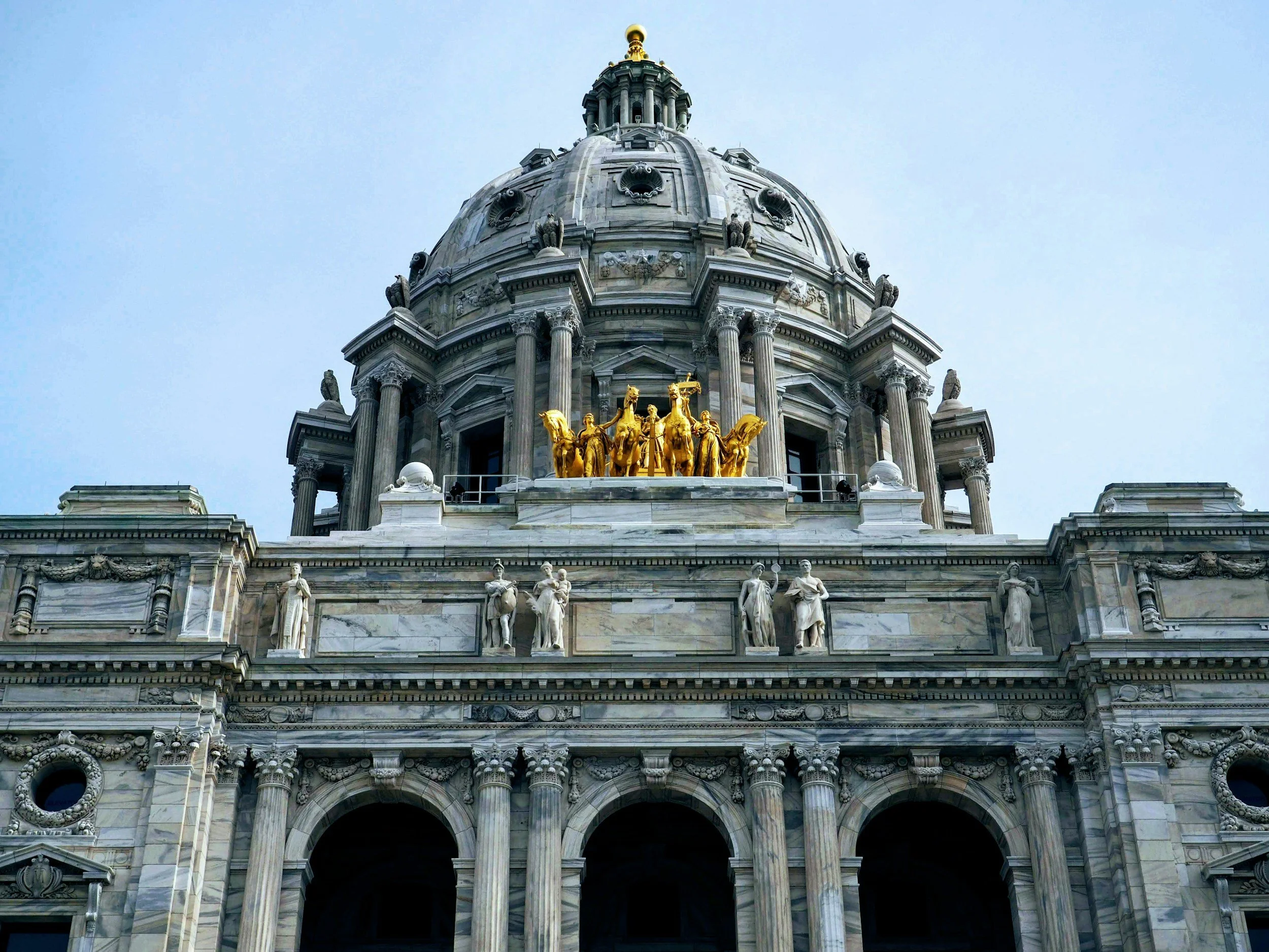 The image shows the top portion of a grand, historic building with a large central dome, adorned with statues and gold accents, against a light blue sky.