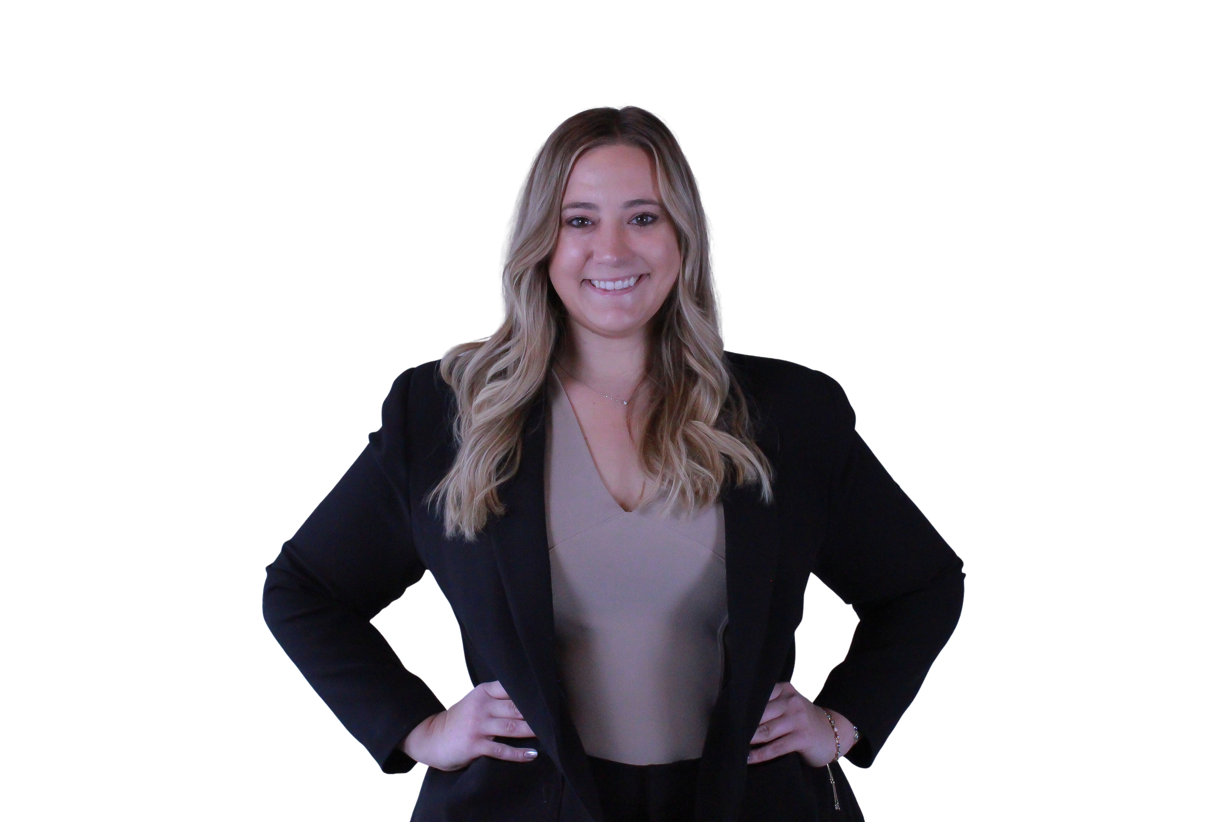 A woman with long wavy blonde hair, wearing a black blazer over a nude-colored top, smiling, standing with hands on hips against a black background.