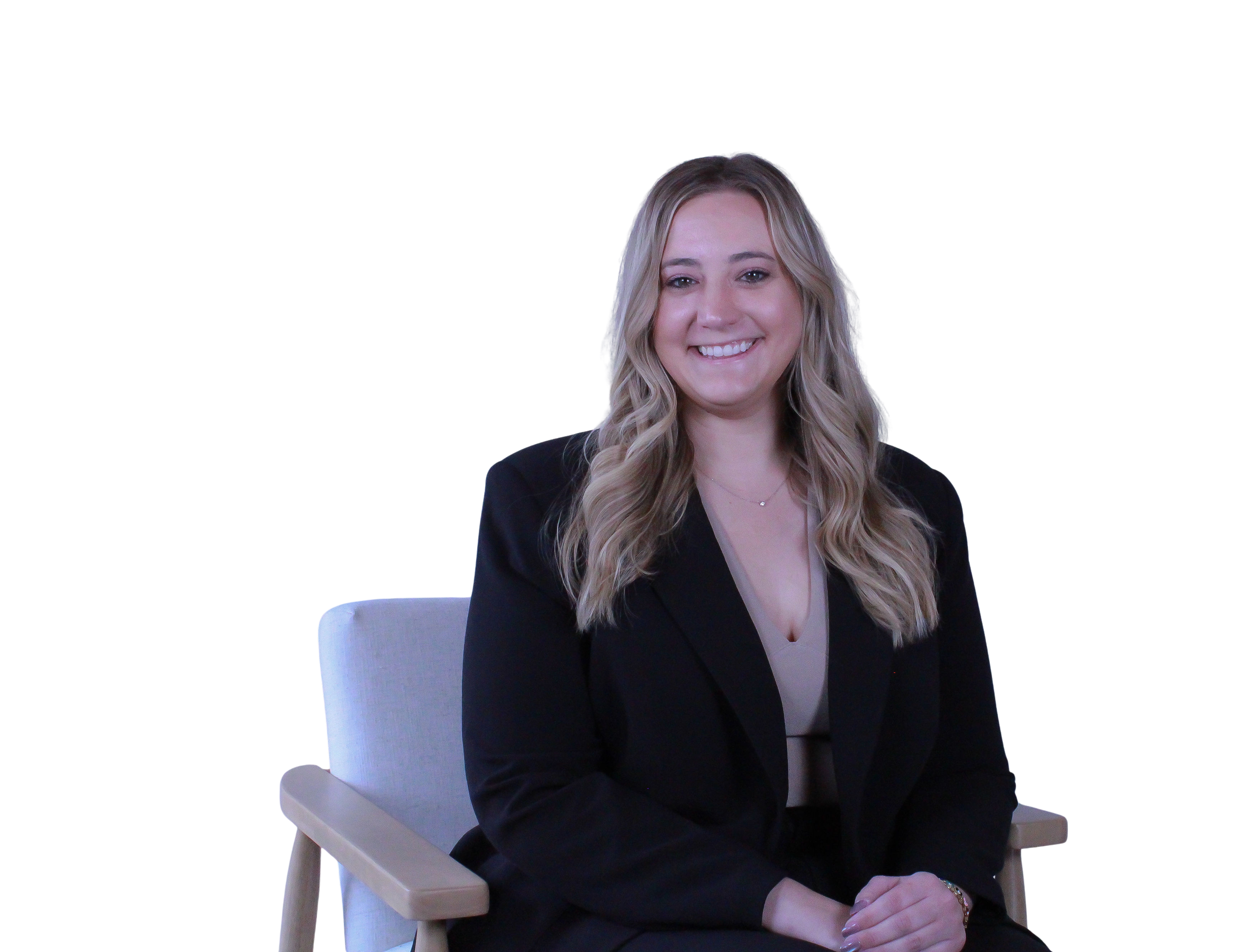 A woman with long blonde hair, smiling, wearing a black blazer and beige top, sitting on a light-colored chair against a black background.
