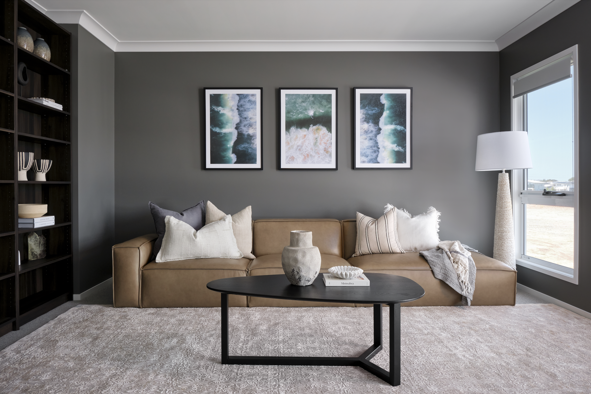 Living room with gray walls, beige sofa with multiple pillows, black coffee table with decorative items, dark bookshelf, and large window with a view outside.