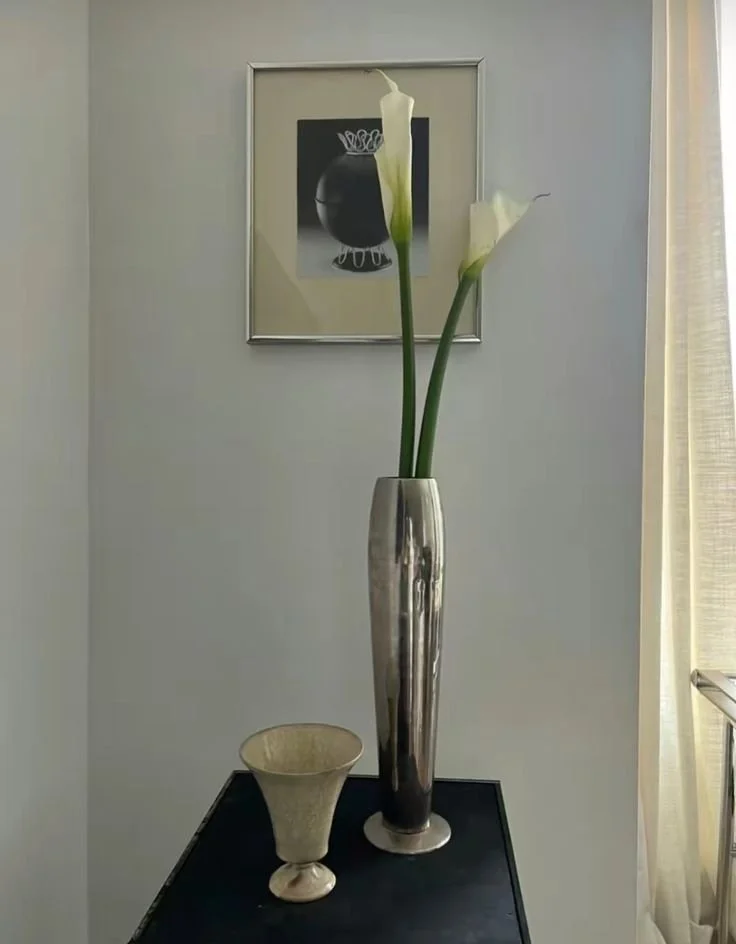A tall, shiny silver vase with three white calla lilies on a black table. A beige bowl is next to the vase. A framed black and white artwork hangs on the light-colored wall behind it, and a window with light curtains is on the right side.