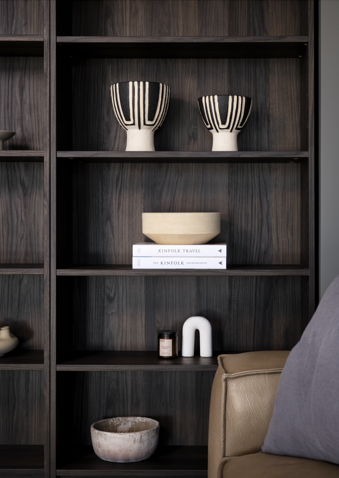 Dark wooden bookshelf with decorative items including two black and white striped vases on the top shelf, books, a beige bowl, a small black jar, a white ceramic U-shaped sculpture, and a large beige bowl on lower shelves, next to a beige sofa with a gray pillow.