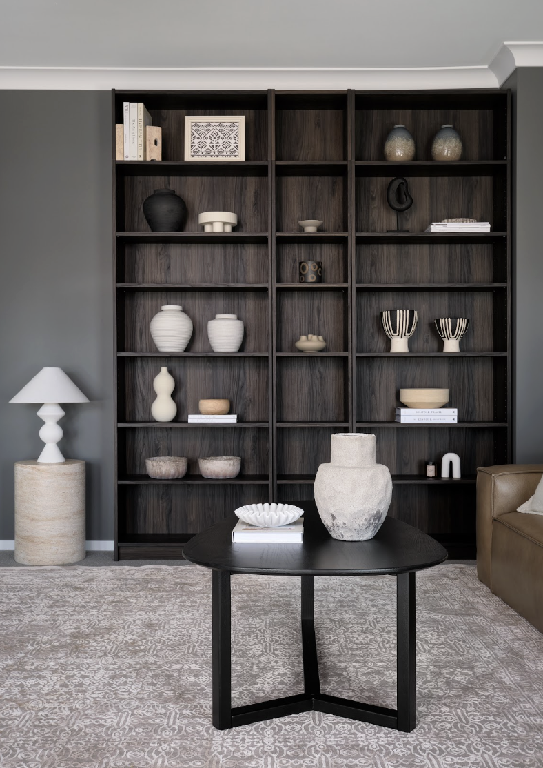 Living room with a dark wood bookshelf filled with various vases, bowls, and books, a black round table with a large stone vase and a book on top, a beige and white table lamp on a stone side table, a brown leather sofa, and a patterned beige rug.