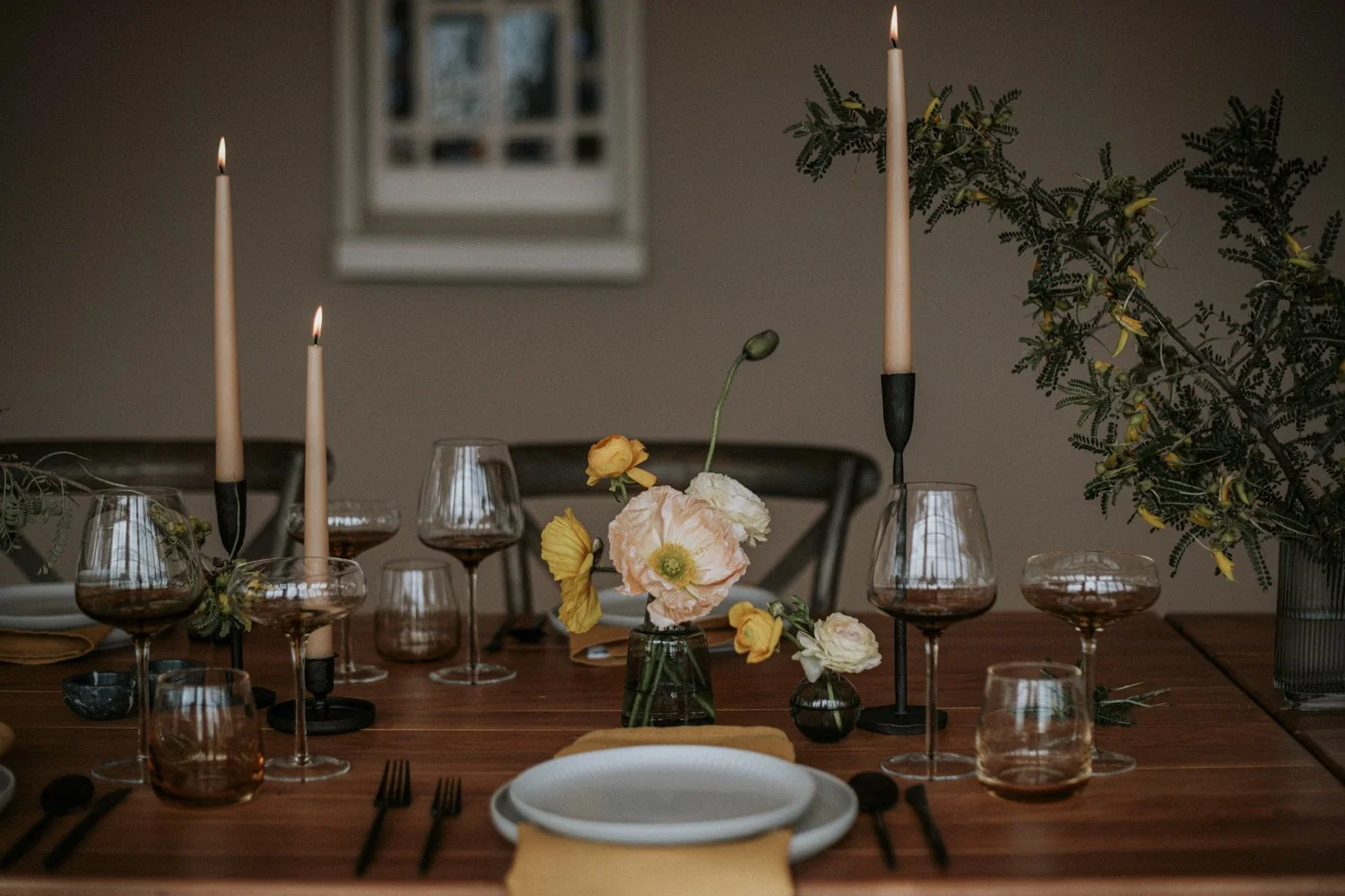 A dining table decorated with candles, flowers, and glassware, set for a meal with plates, utensils, and napkins, in a warmly lit room with a window in the background.