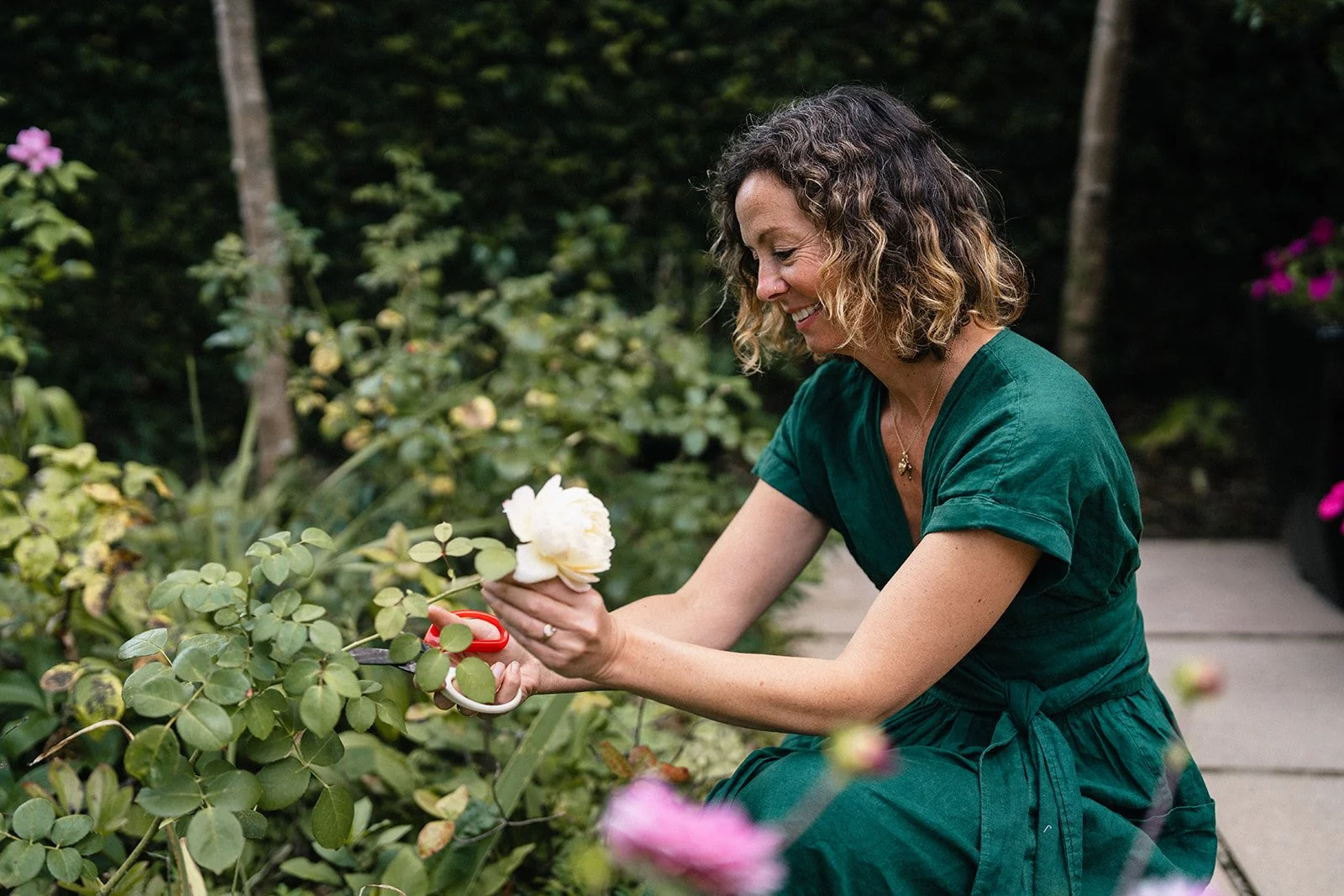 A woman in a green dress is trimming a bush with pruning shears and holding a flower.