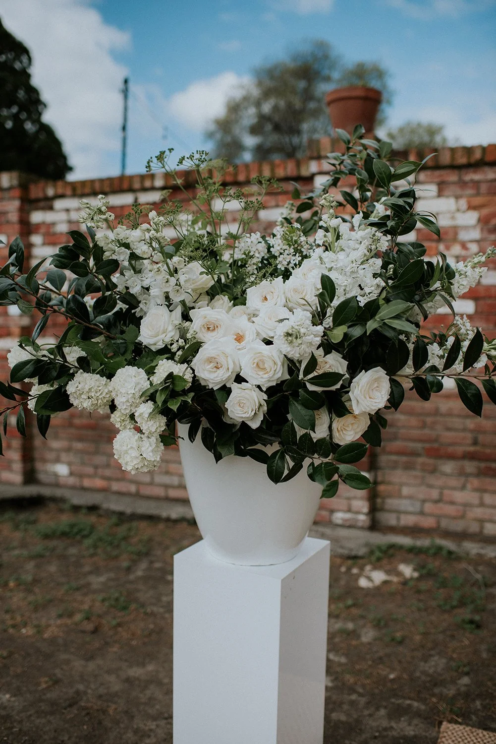 White wedding flower arrangement in a white urn on a white plinth, with a red brick wall and blue sky in the background.