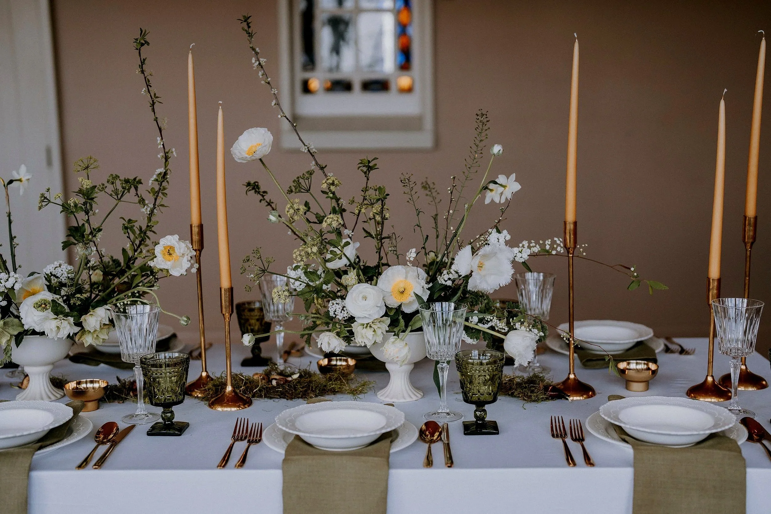 A decorated dining table with white and green floral centerpieces, spring flowers including ranunculus, poppies and blossom, gold and green glassware, white plates with gold trim, gold utensils, and beige napkins. The table is set for a wedding.