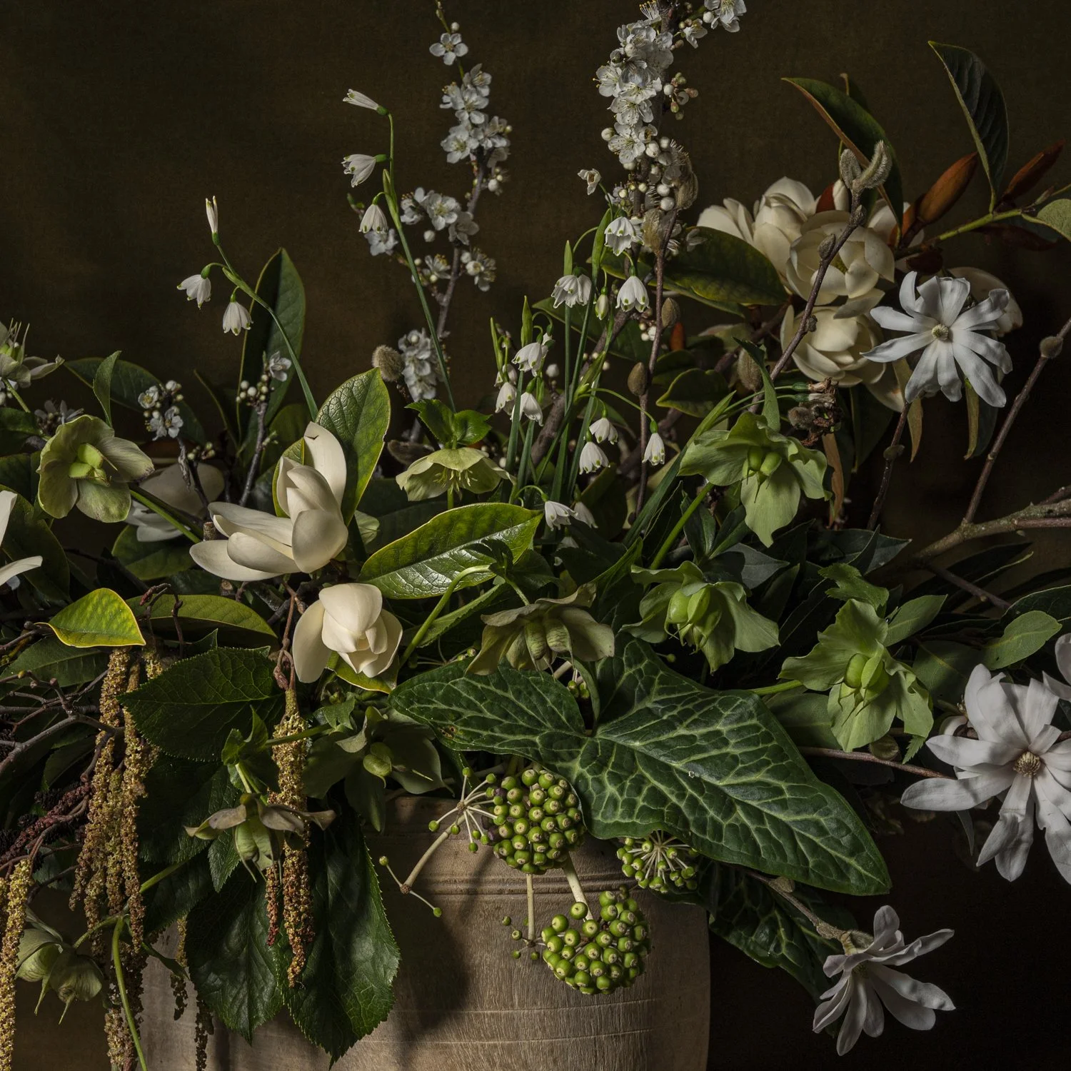 A floral arrangement with white flowers, green leaves, and berries in a beige vase against a dark background.