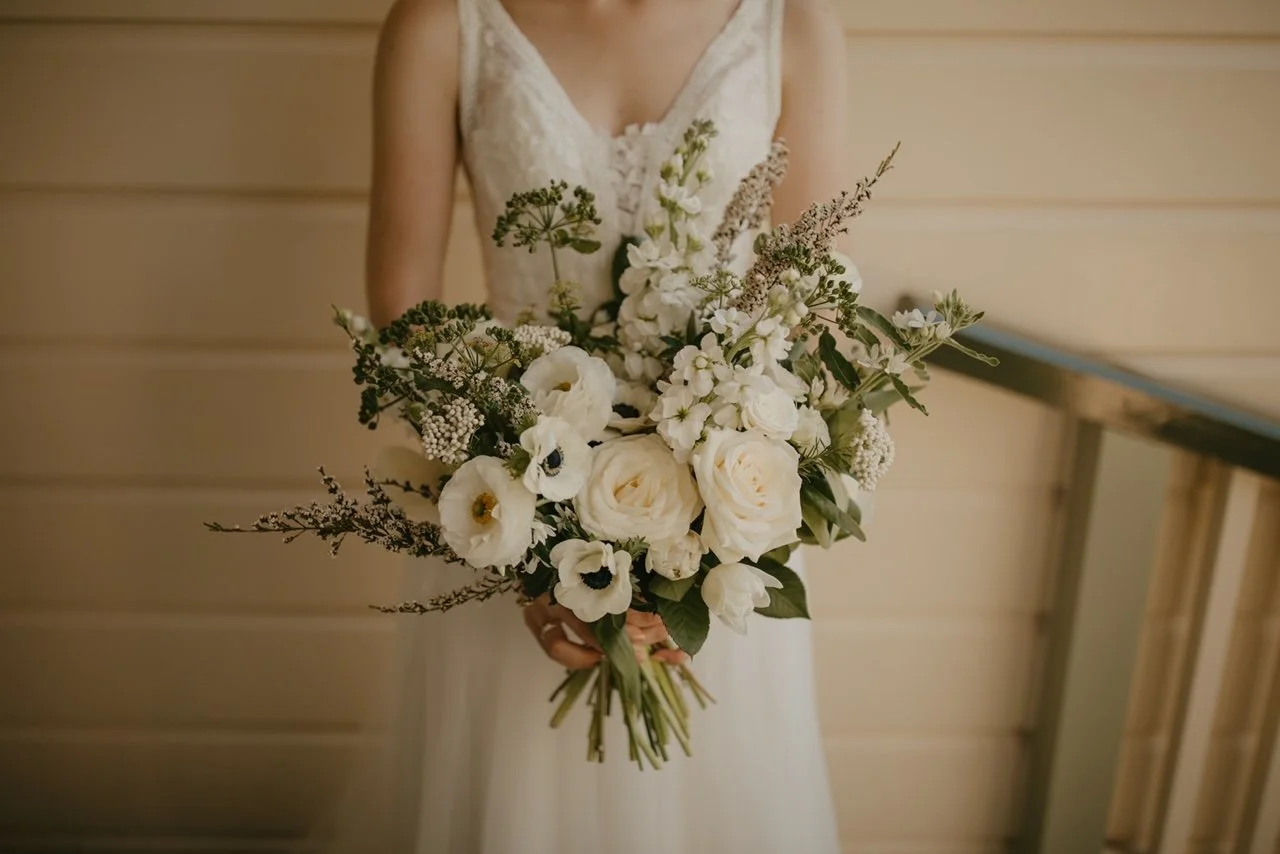 Bride holding a bouquet of white and cream-colored flowers, including roses, anemones, and greenery, in front of a wooden wall.