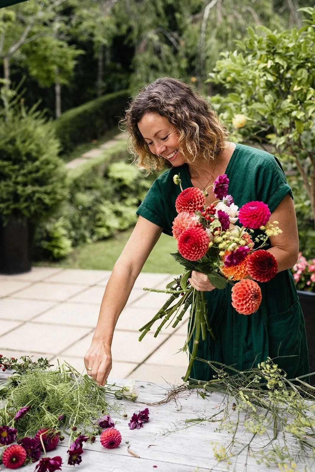 A woman in a green dress arranging a colorful flower bouquet outdoors on a wooden table, surrounded by green plants and trees.