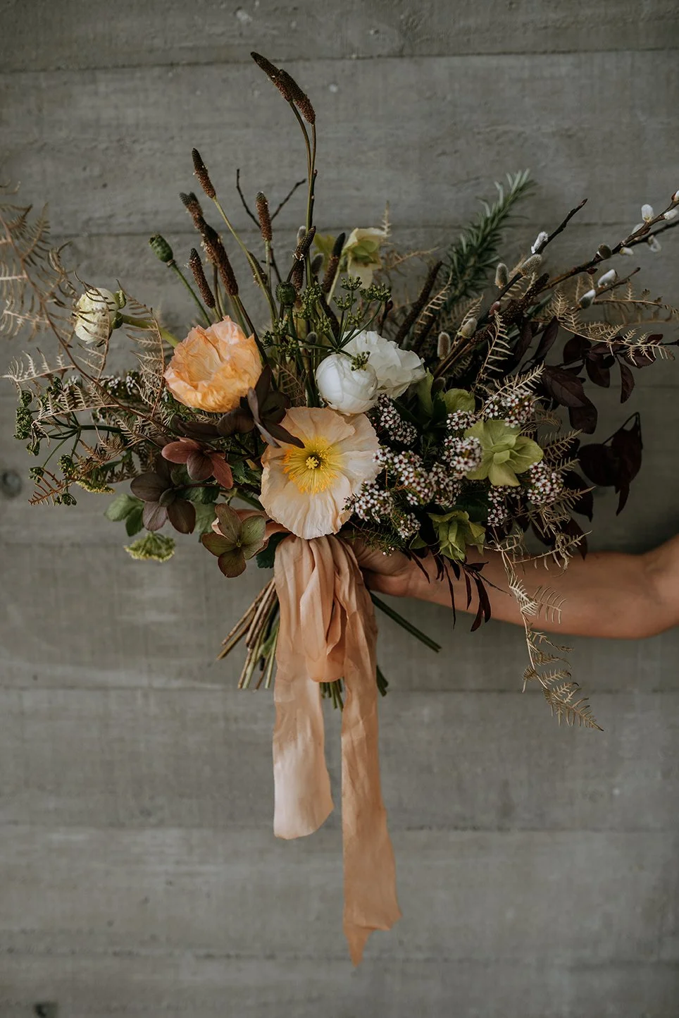 A bridal bouquet of spring flowers and muted brown foliage with a peach-colored silk ribbon against a concrete background.
