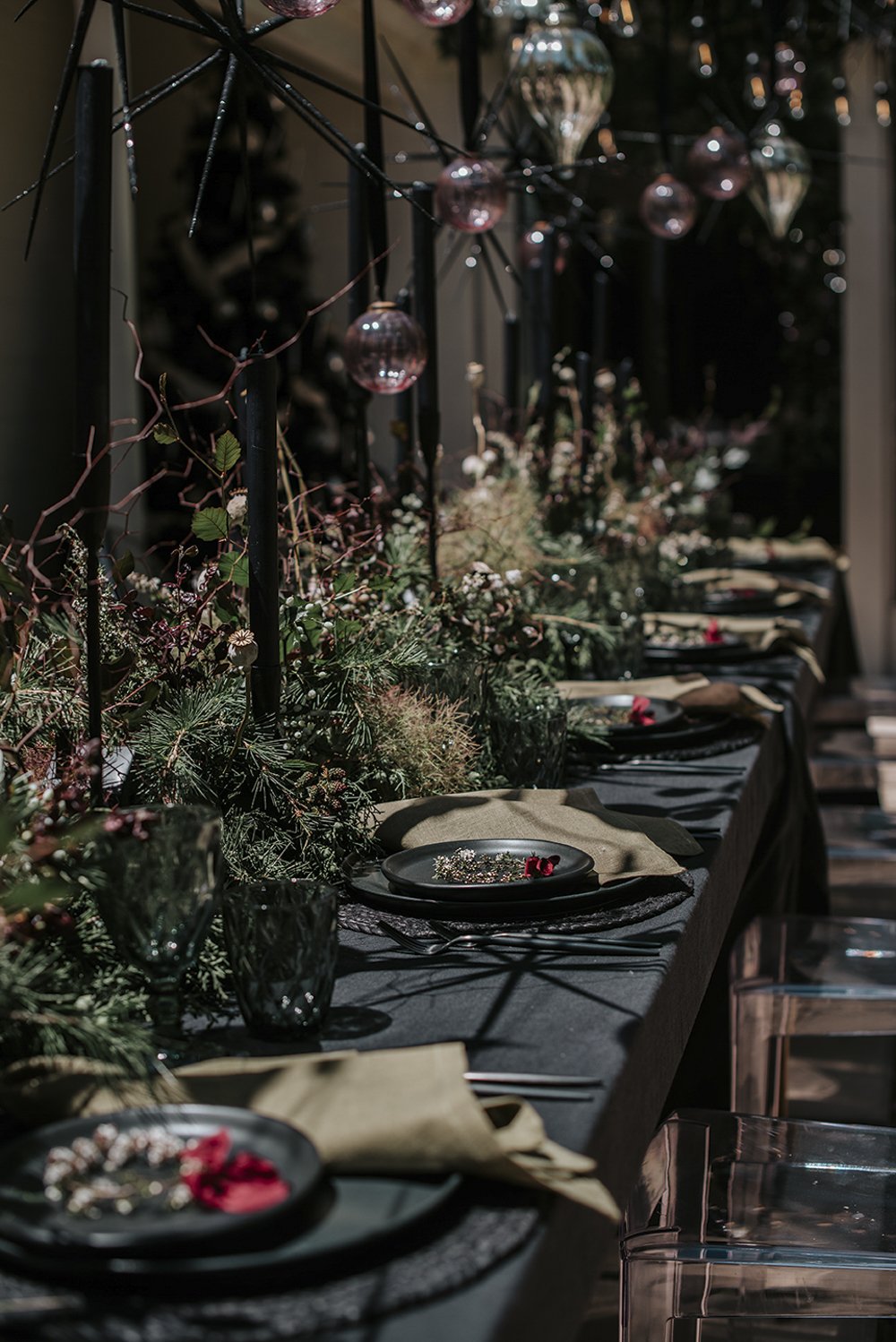 Elegant dining table decorated with greenery, black plates, napkins, and glassware, with hanging glass ornaments and dim lighting.
