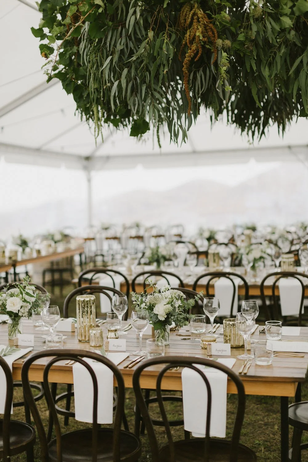 Elegant wedding reception with neutral flower centrepiece's on wooden tables in marquee with a large green suspended foliage installation