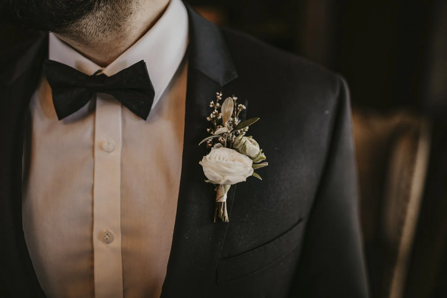 Close-up of a groom in a black tuxedo, white dress shirt, and black bow tie, with a boutonniere of white ranunculus and greenery pinned to his lapel.