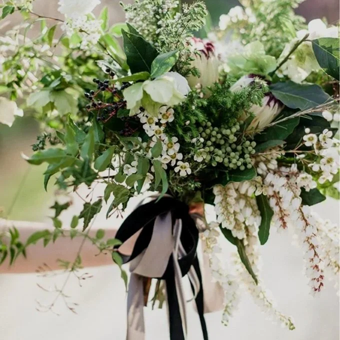 A close-up of a bridal bouquet with white and green flowers, including hellebores, protea, pieris, foliage, and a black and white ribbon.