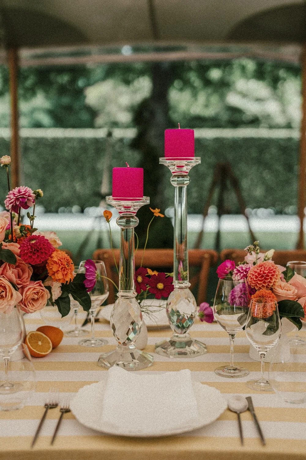 A table decorated with pink candles on glass candlesticks, bright summery floral arrangements in vases, yellow and white striped tablecloth, citrus decoration on table, set for a celebration or dinner event.