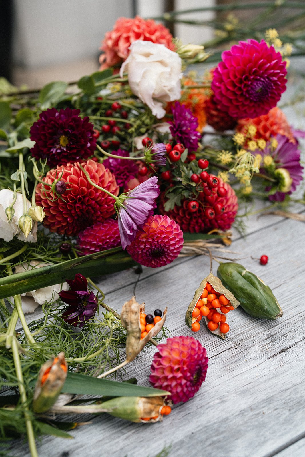 A colorful assortment of fresh flowers and berries on a wooden surface, including dahlias, roses, and berries, some of which are spilling out of seed pods.