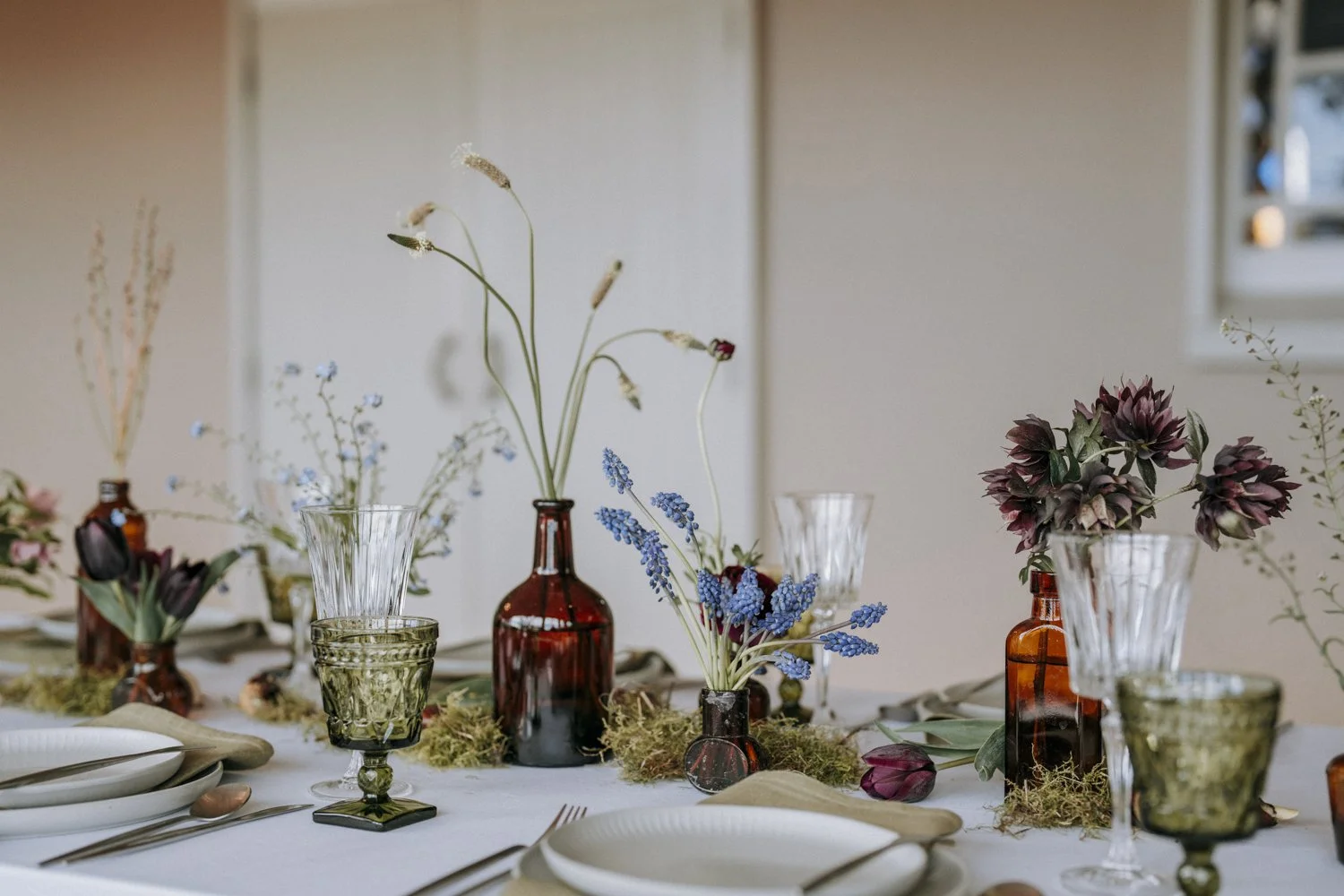 Table setting with moss and flowers in brown glass bottles including grape hyacinth, tulips, hellebores and seed heads