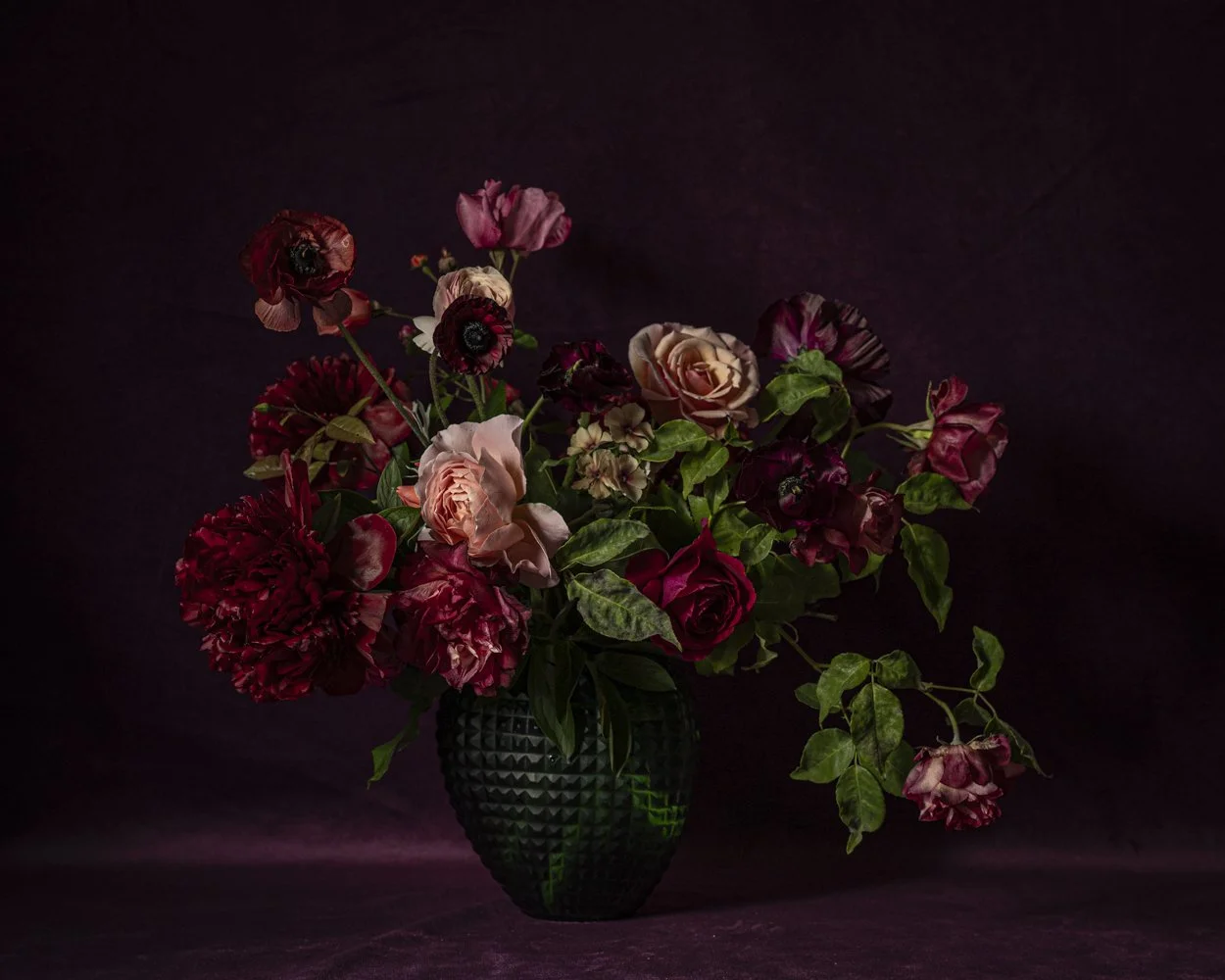 A dark green textured vase holding a mixed bouquet of pink, red, and purple flowers, against a dark background.