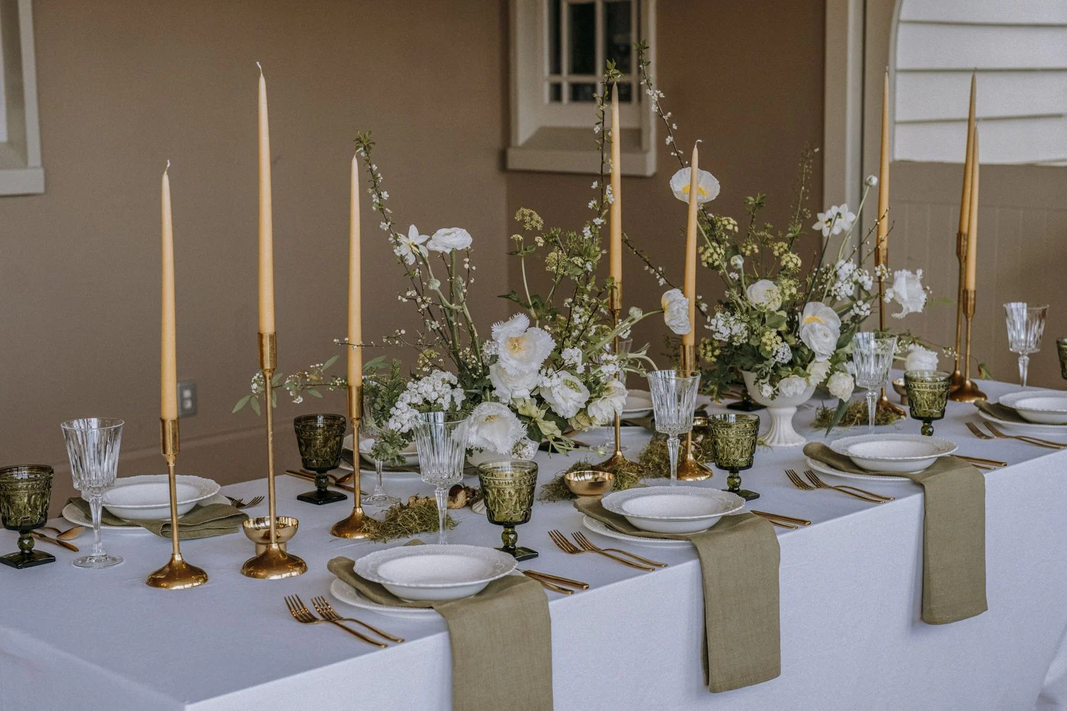A decorated indoor dining table with white tablecloth, gold and green accents, and floral centerpieces with white flowers, tall gold candlesticks, and various glassware and dinnerware.