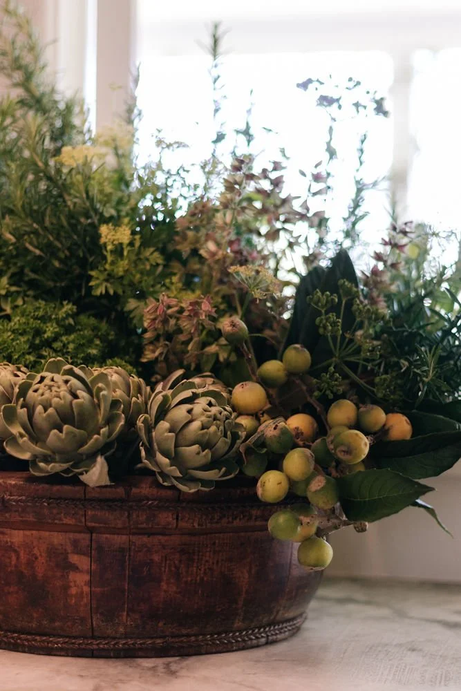 A round wooden planter filled with various green plants, including artichokes, loquat, and herbs, placed on a light-coloured surface near a window with bright natural light.