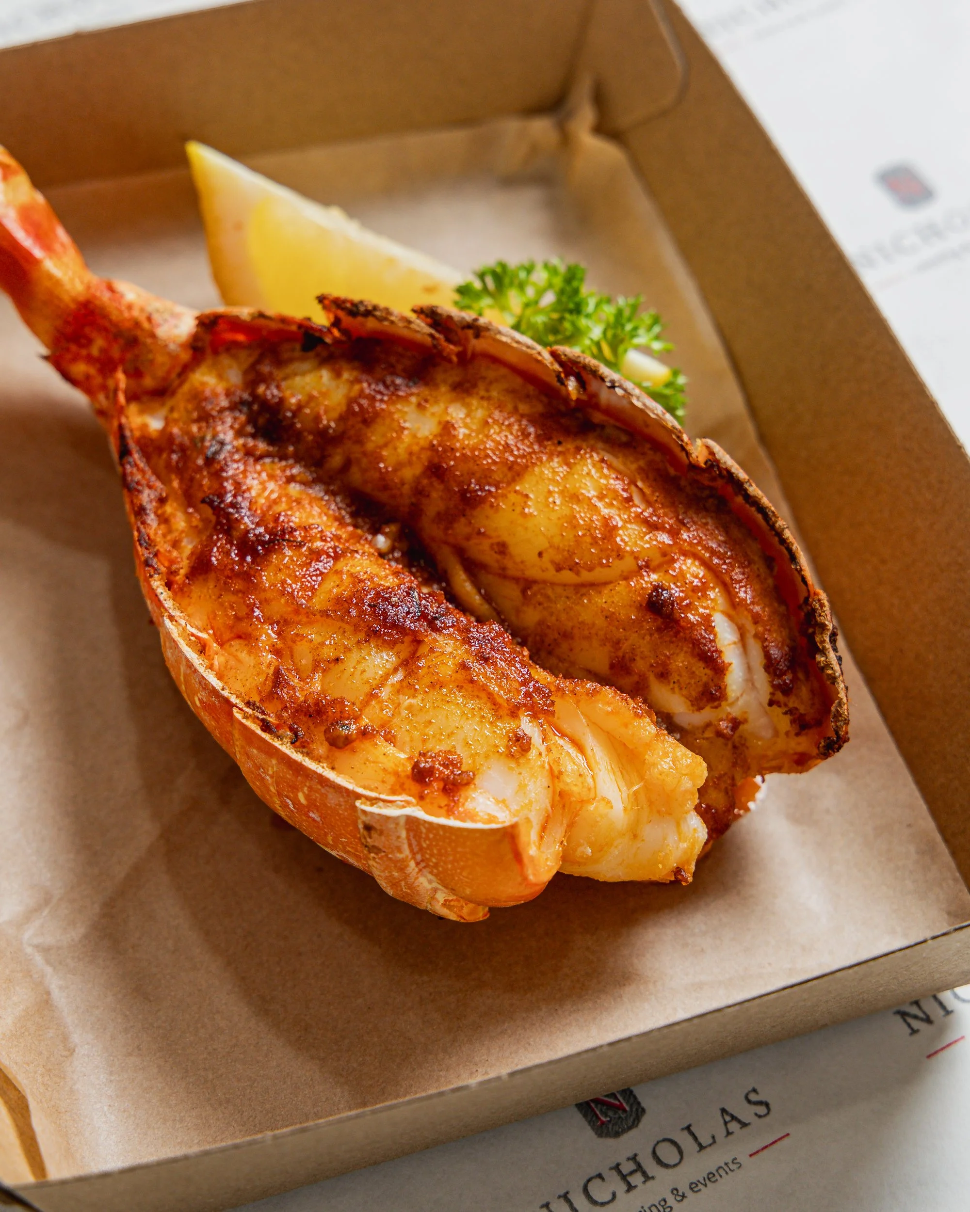 Lobster tail with grilled seasoning, lemon wedge, and parsley garnish in a brown box on a paper plate.
