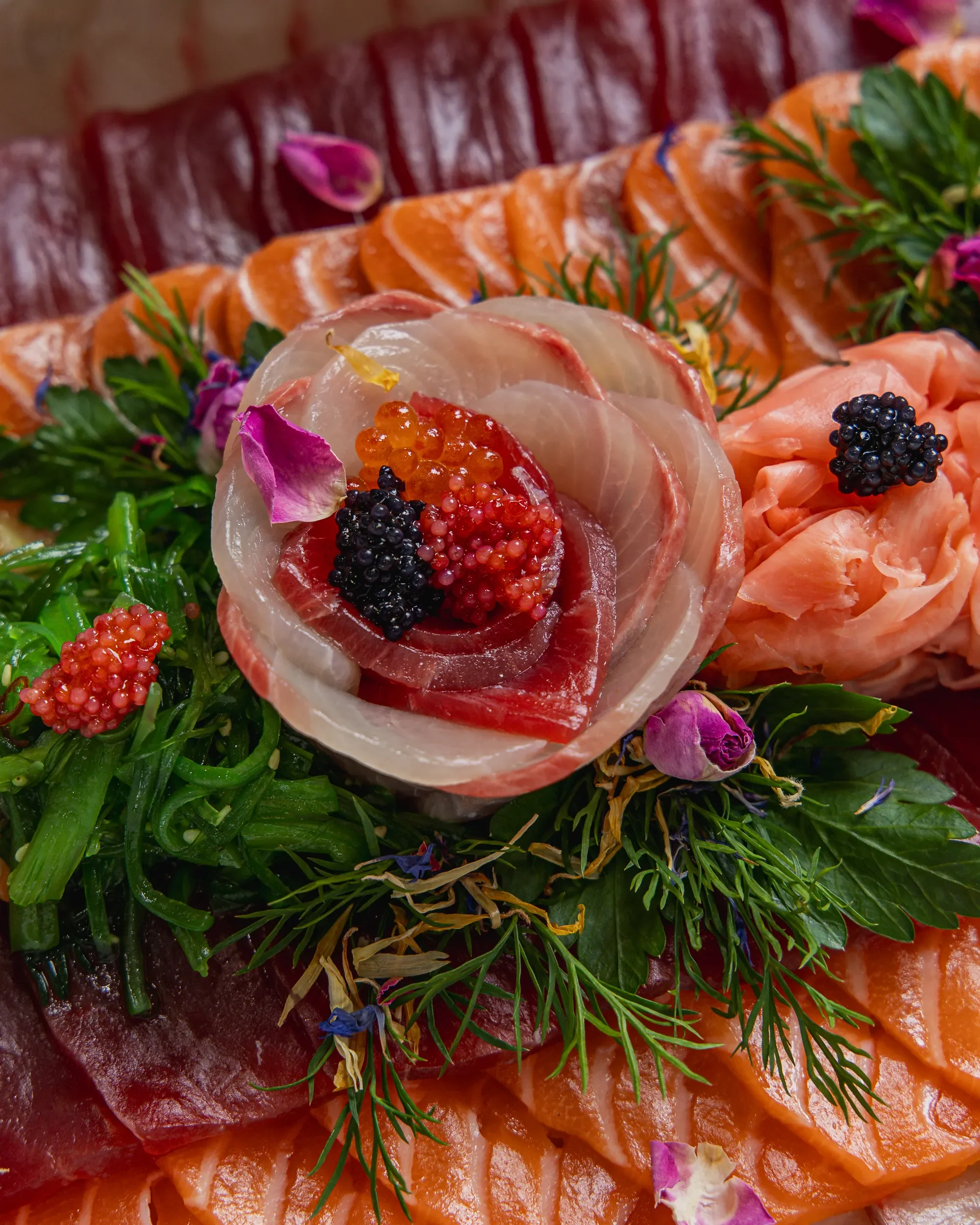 Assorted sashimi including slices of salmon, tuna, and white fish, garnished with edible flowers and fish roe, on a bed of greens.