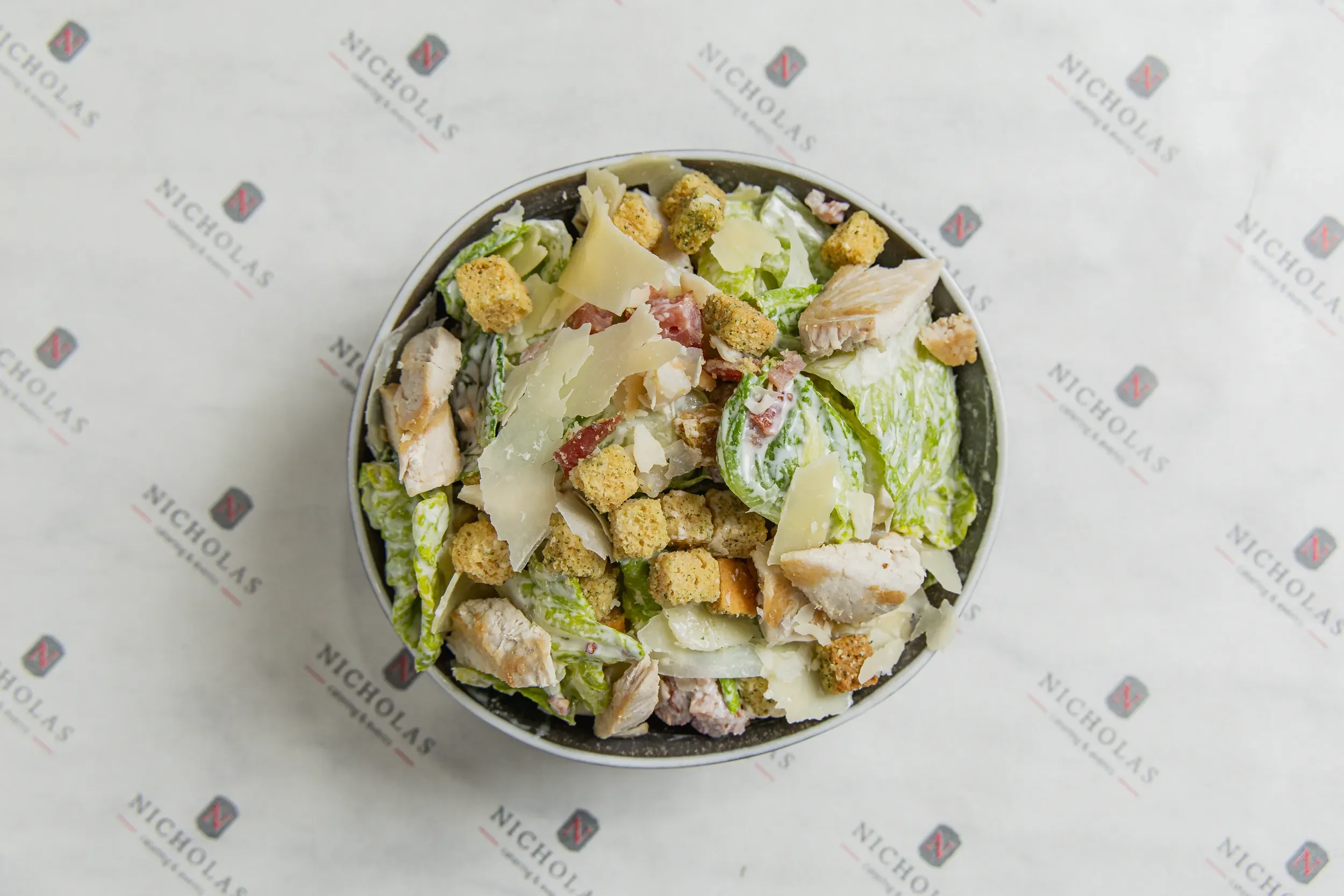 Top view of a Caesar salad in a bowl with lettuce, grilled chicken pieces, parmesan cheese, croutons, and bacon bits, on branded paper.